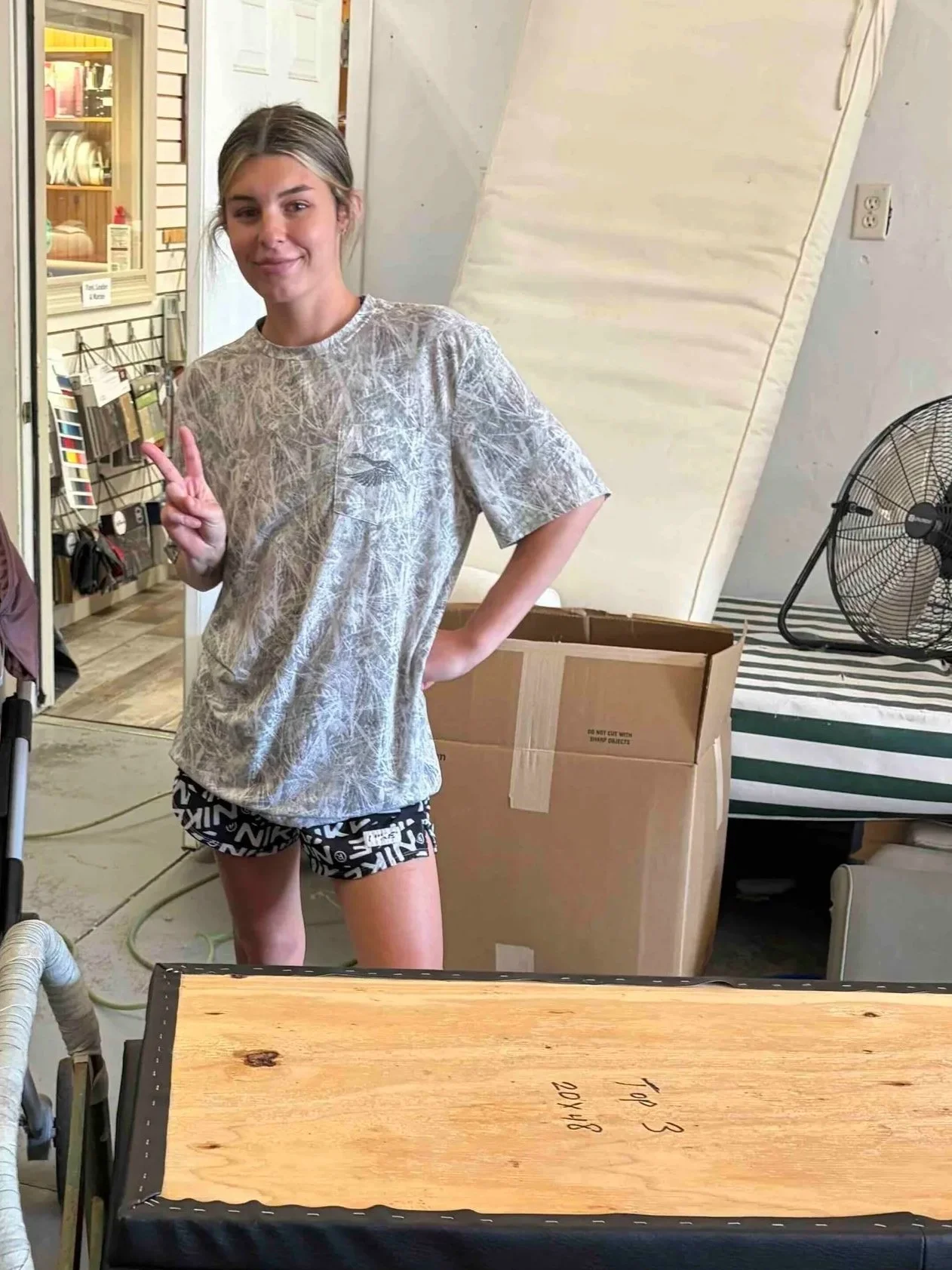Young woman smiling and making a peace sign pose inside a store, wearing a gray patterned T-shirt and black shorts, with a cardboard box, a fan, and various store items around her.