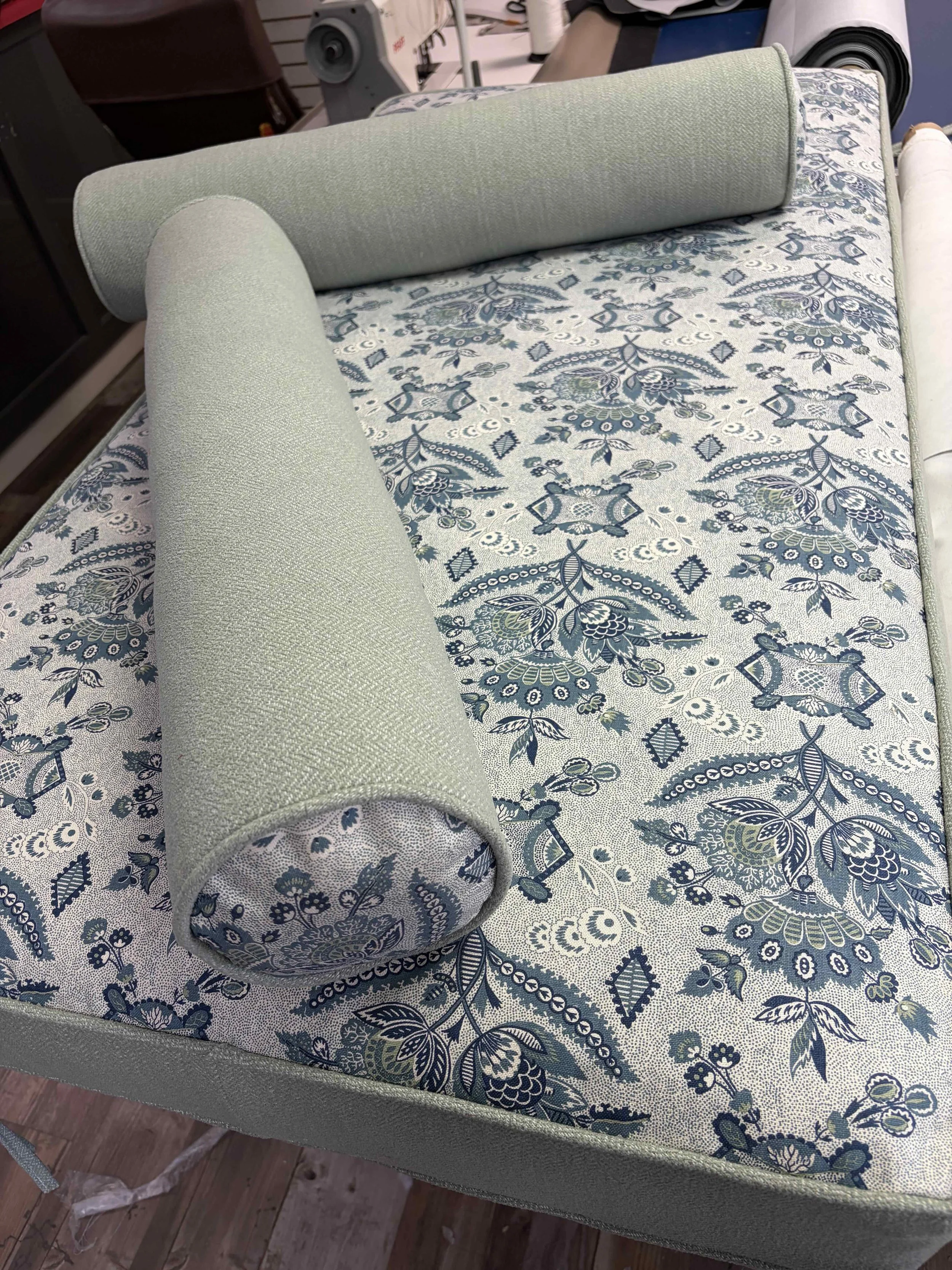 A section of fabric with a floral pattern in shades of blue, green, and white, partially rolled over a padded surface, likely a piece of furniture or upholstery.
