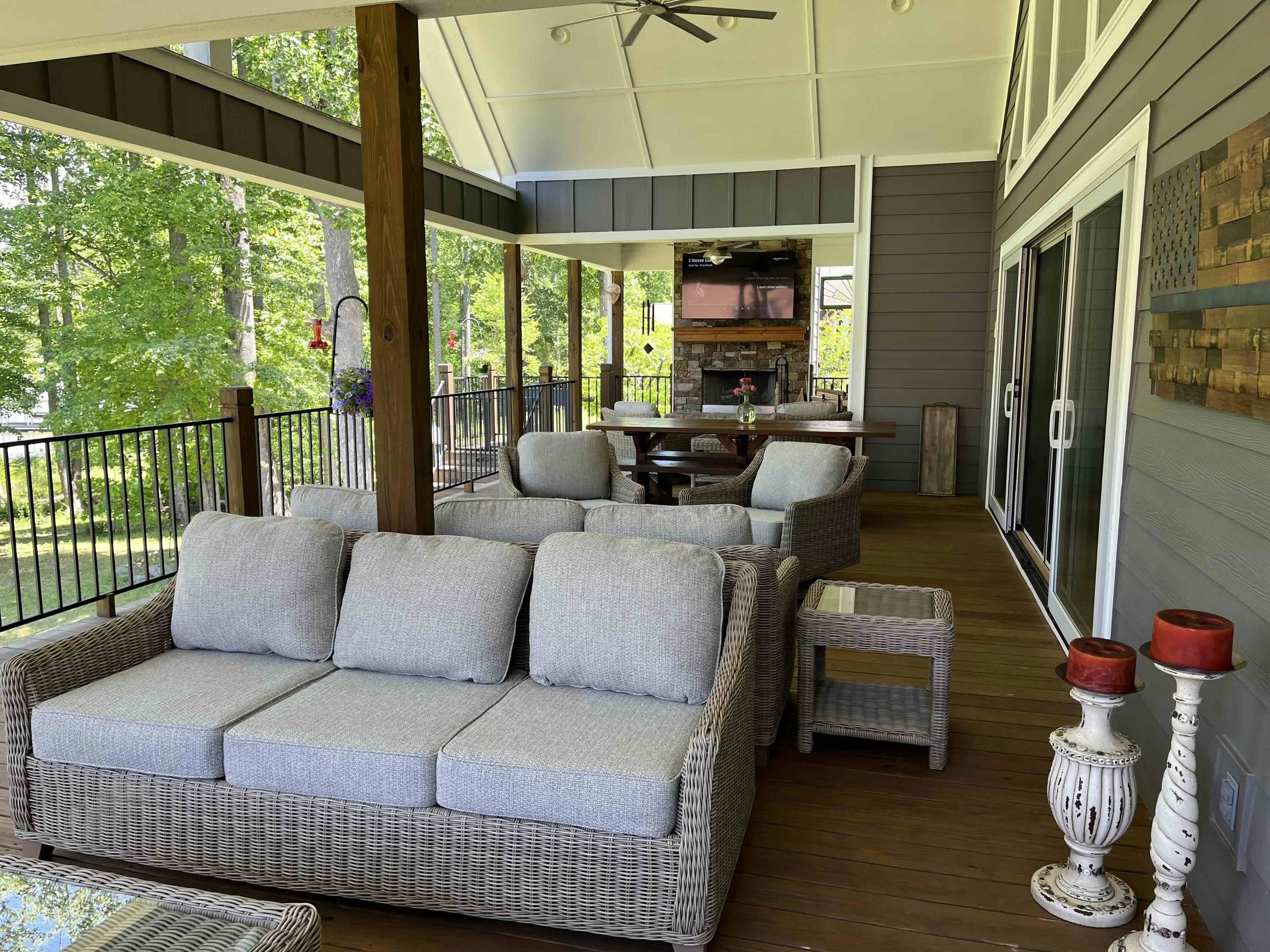 An outdoor covered porch with wicker furniture, including a sectional sofa with light-colored cushions, and a round wooden table with four chairs. The porch has a ceiling fan, a large flat-screen TV mounted above a stone fireplace, and decorative elements like red candles on a white side table and hanging flower baskets. The porch is surrounded by a black metal railing and overlooks lush green trees.
