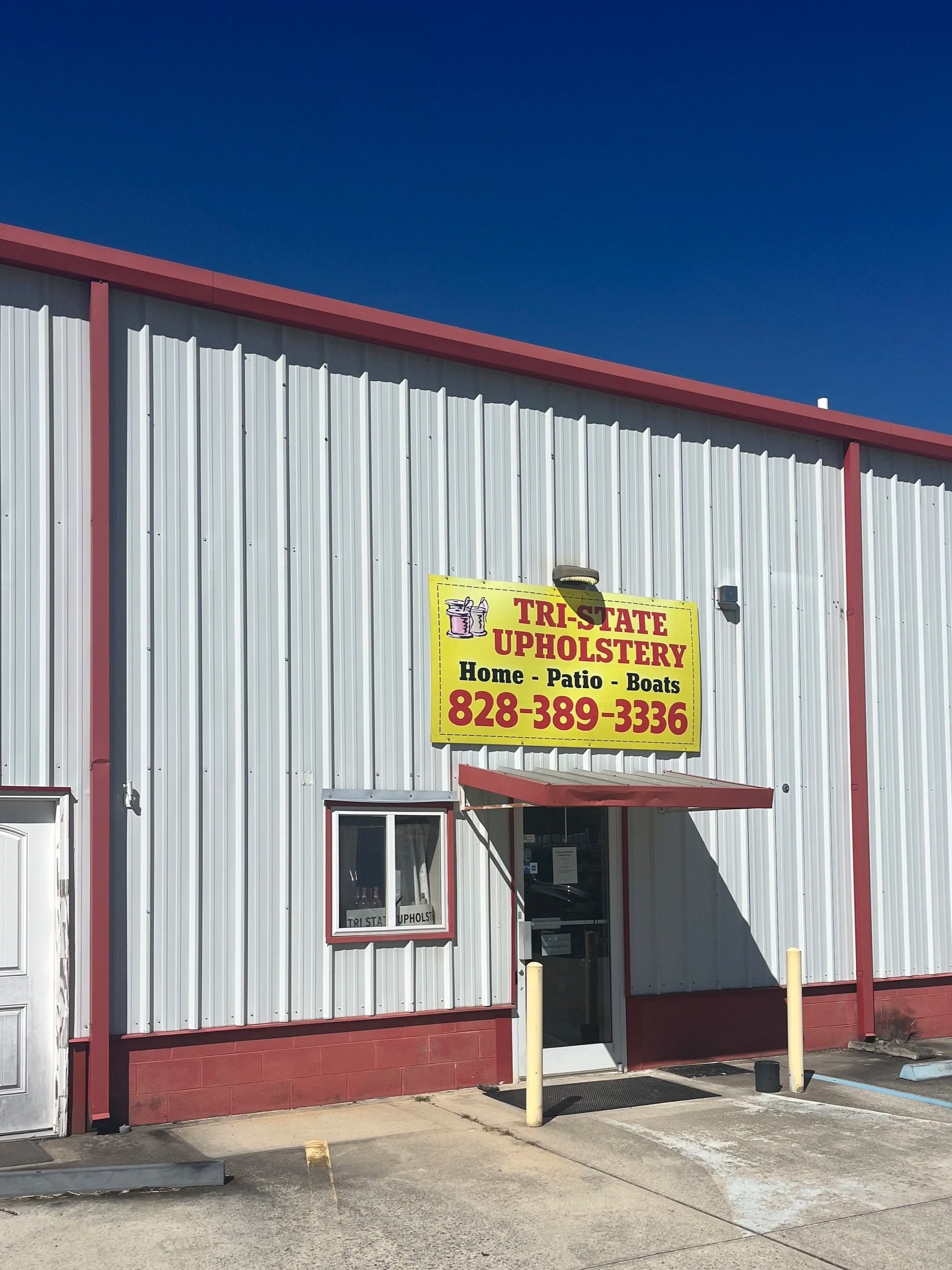 Exterior of a metal building with a yellow sign that reads 'Tri-State Upholstery' and lists services like home, patio, and boats, with a phone number. The building has a small window and entrance door, with parking spots in front.