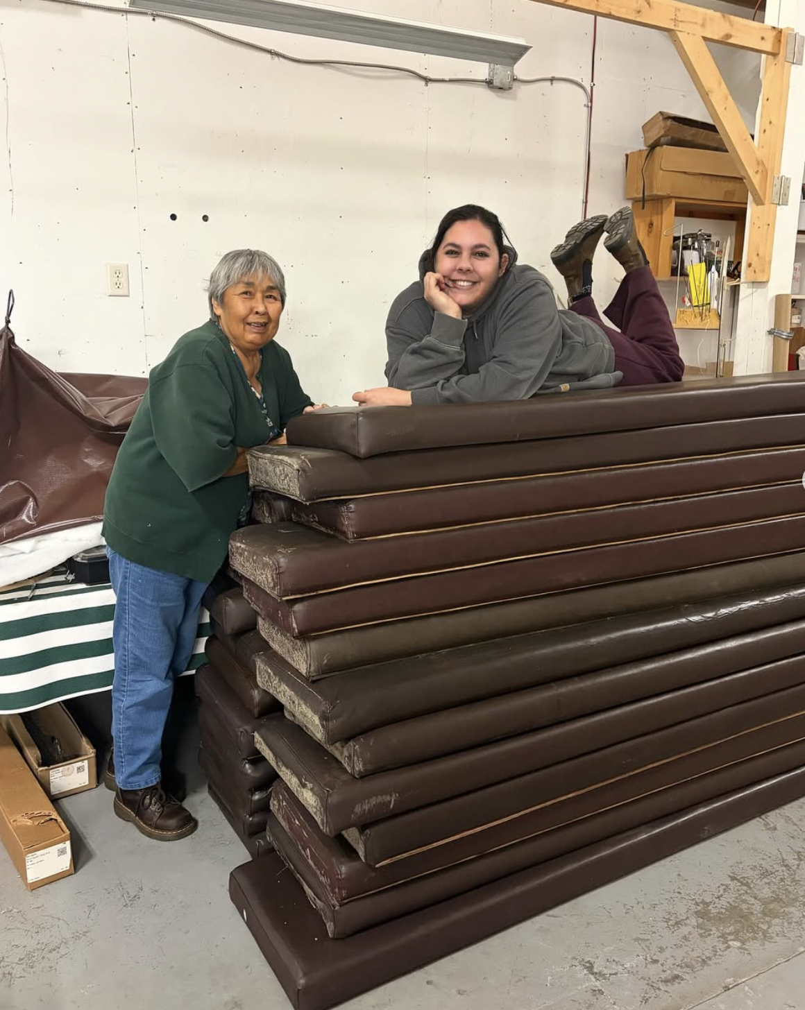 Two women, one older and one younger, posing with a stack of brown couches in a warehouse.