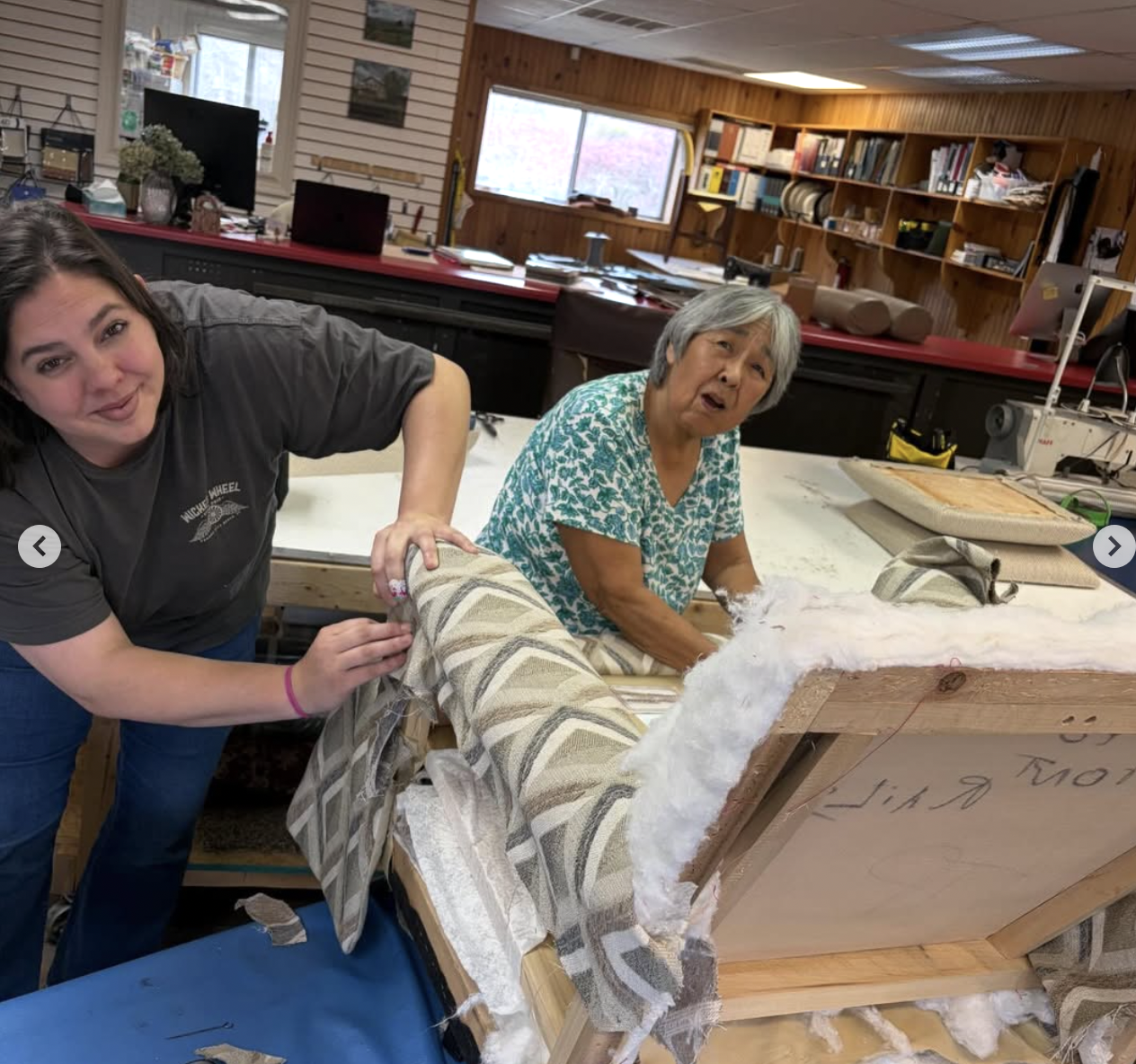Two women working together on an upholstery project in a workshop, with fabric and foam on a wooden frame, surrounded by tools and materials.
