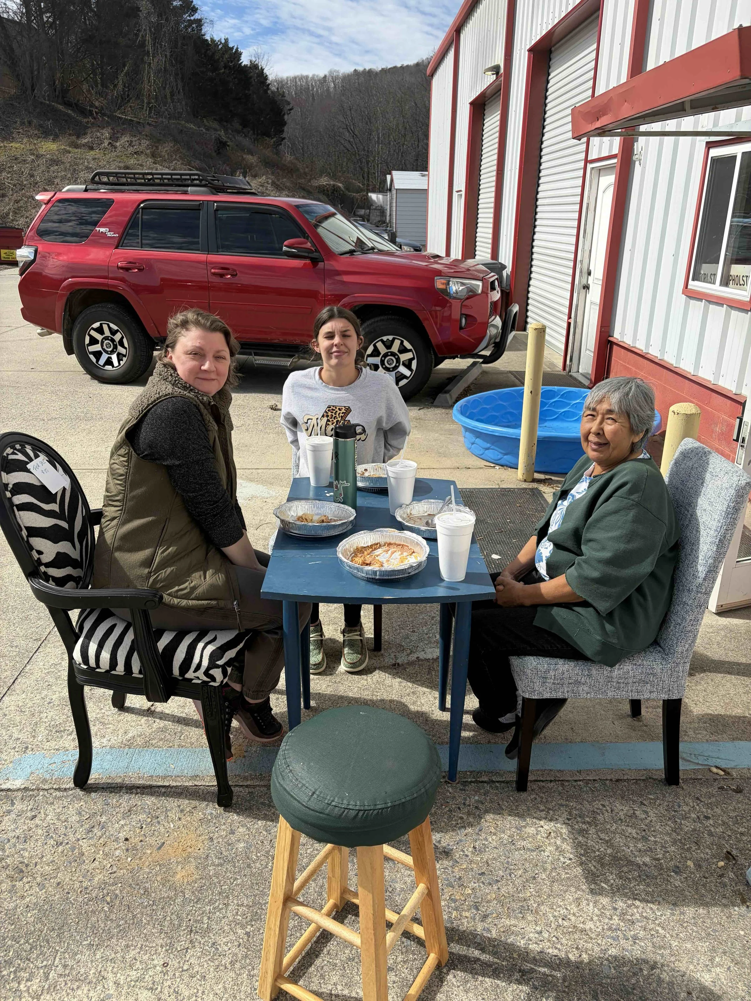 Three women sitting at a table outdoors, eating and smiling. Two of them are seated on chairs, and one is sitting on a cushioned chair. There are food containers and drinks on the table. In the background, there is a red SUV truck parked beside a building with metal siding, a blue plastic kiddie pool, and a landscape with trees and hills.