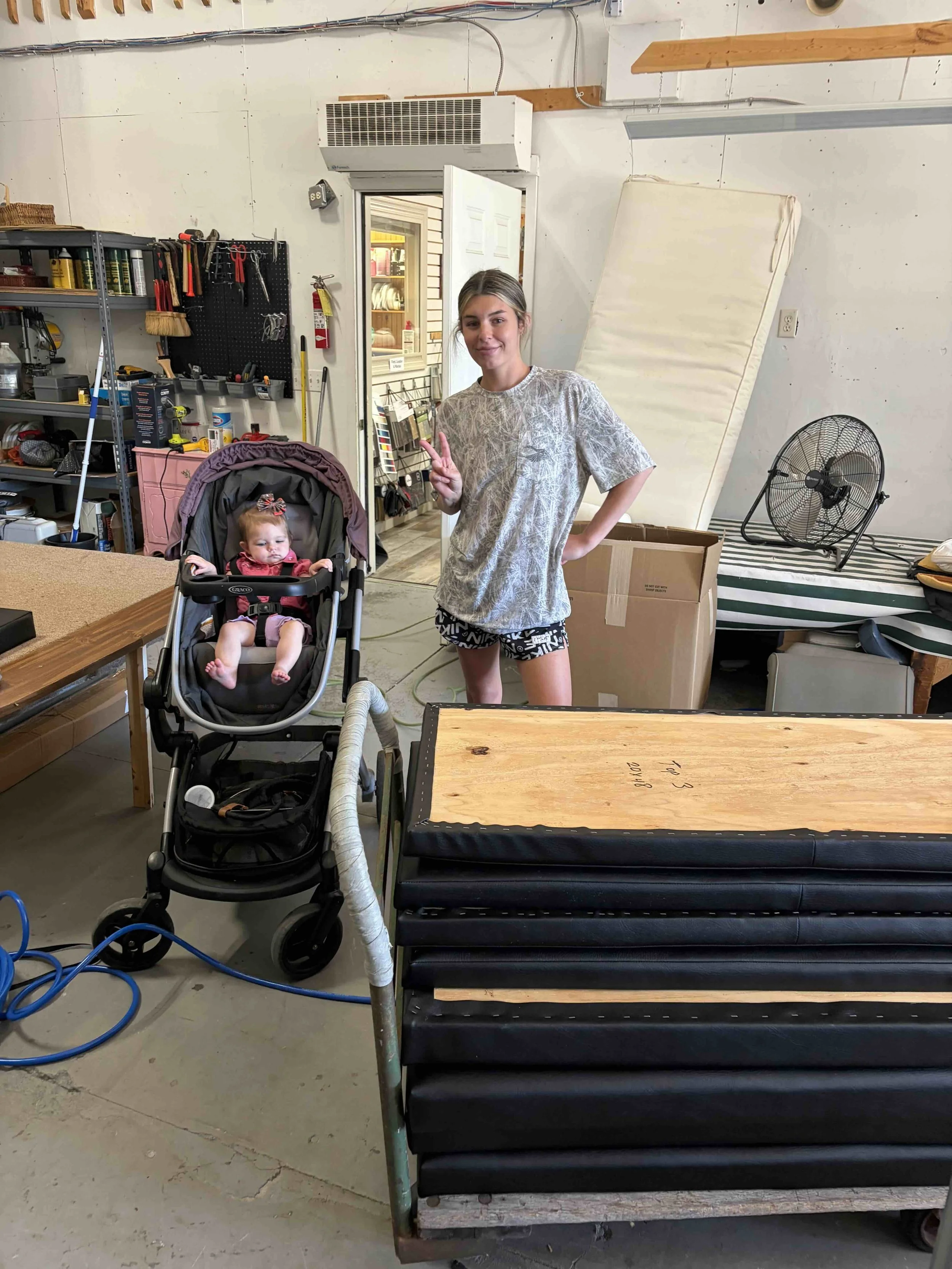 A young woman with gray hair and a patterned shirt standing in a workshop next to a young girl sitting in a stroller. The woman is making a peace sign with her right hand and smiling. The workshop has shelves with tools, a fan, and a large piece of foam or mattress leaning against the wall.