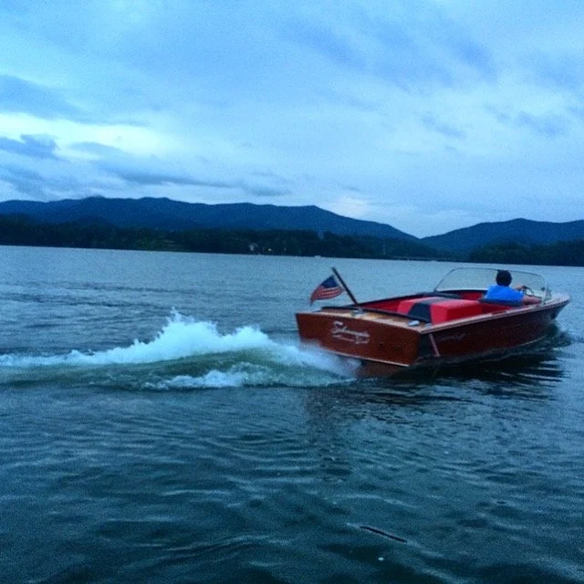 A red and black speedboat with an American flag is moving across a lake with a person sitting in the boat, against a backdrop of mountains and cloudy sky.