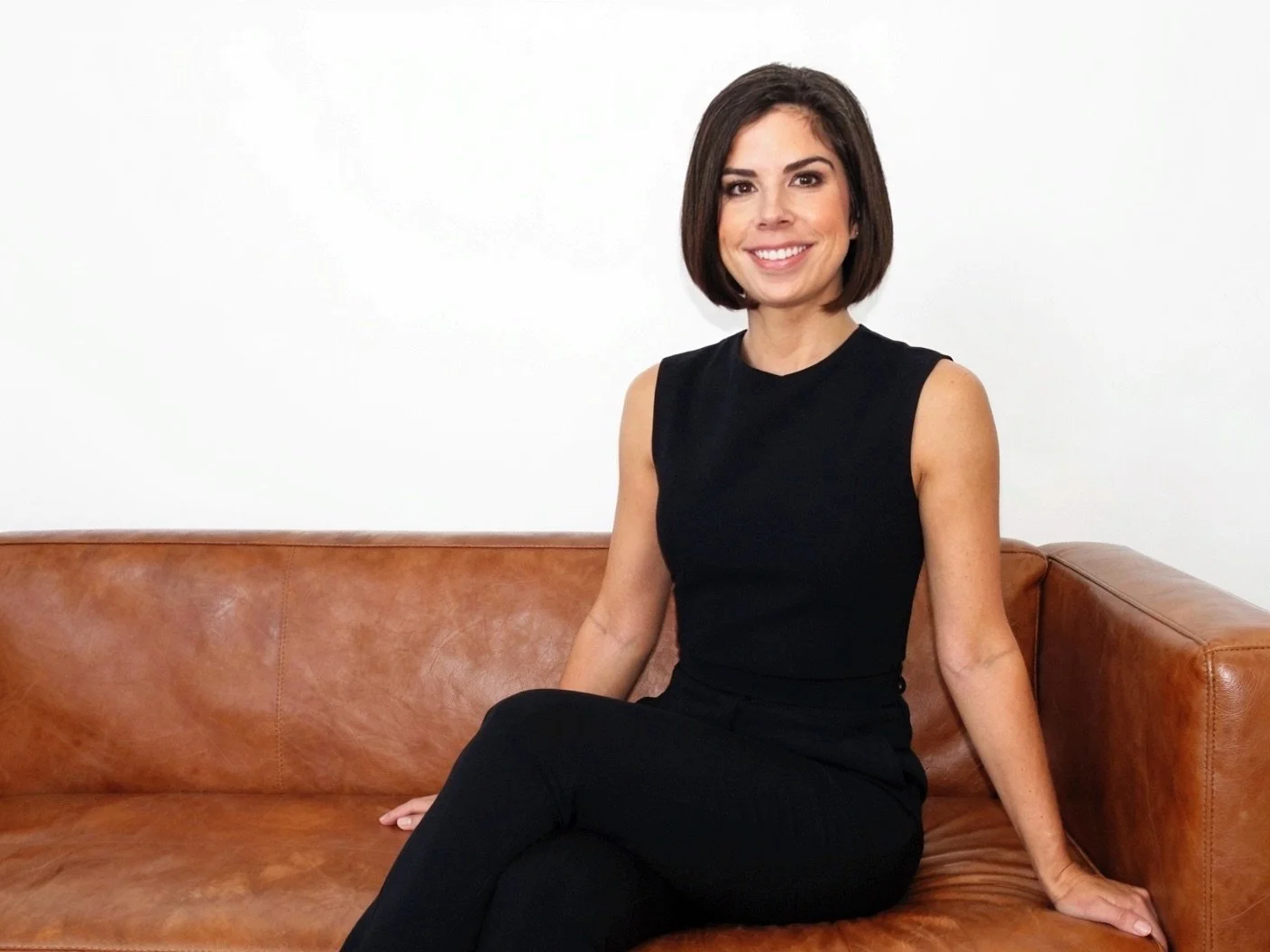 A woman with short dark hair sitting on a brown leather couch against a plain white wall, smiling, wearing a black sleeveless top and black pants.
