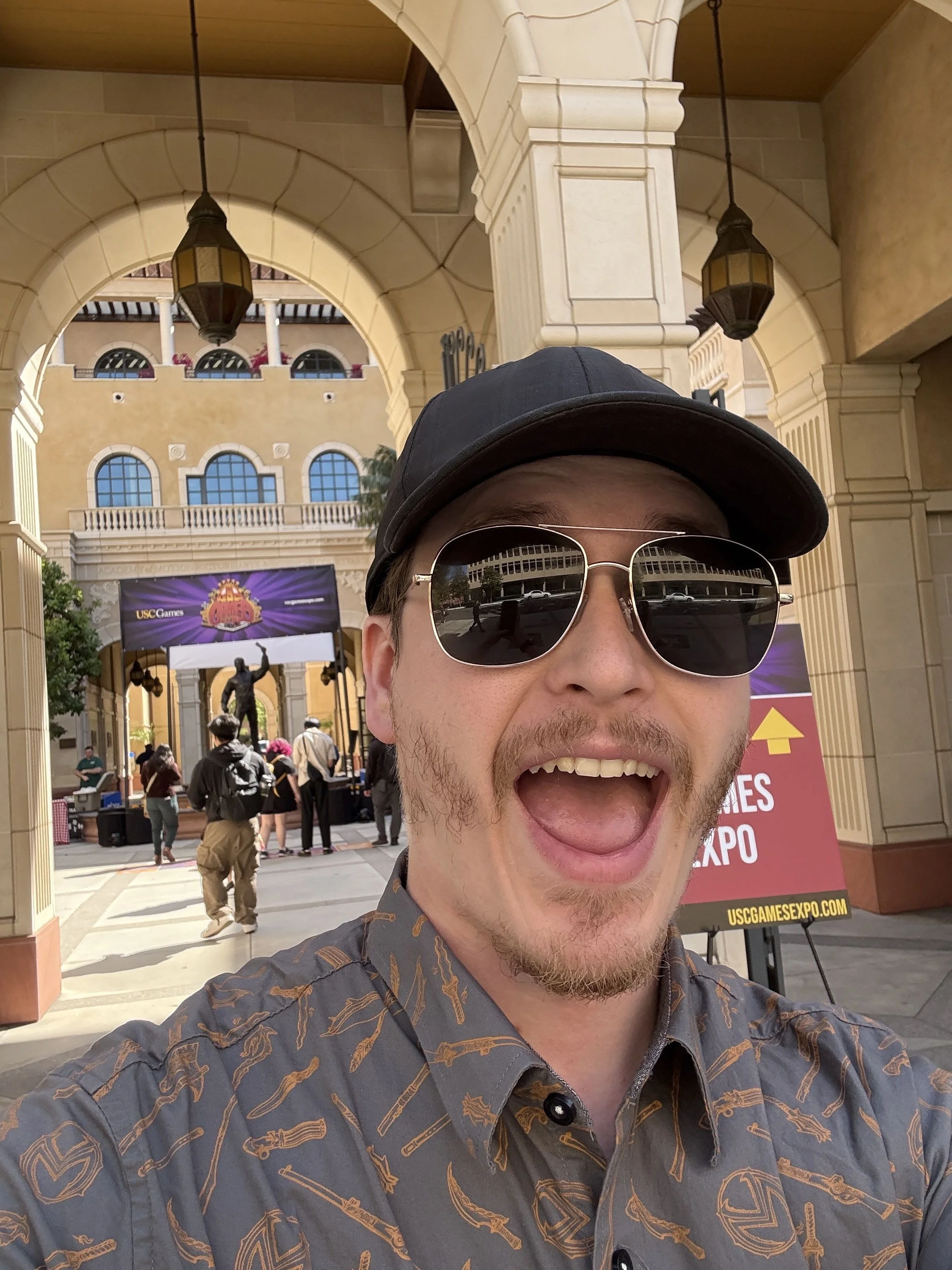 A man wearing sunglasses, a black cap, and a patterned shirt taking a selfie at an outdoor event with a decorative building, people, and a sign for USC Game Expo in the background.