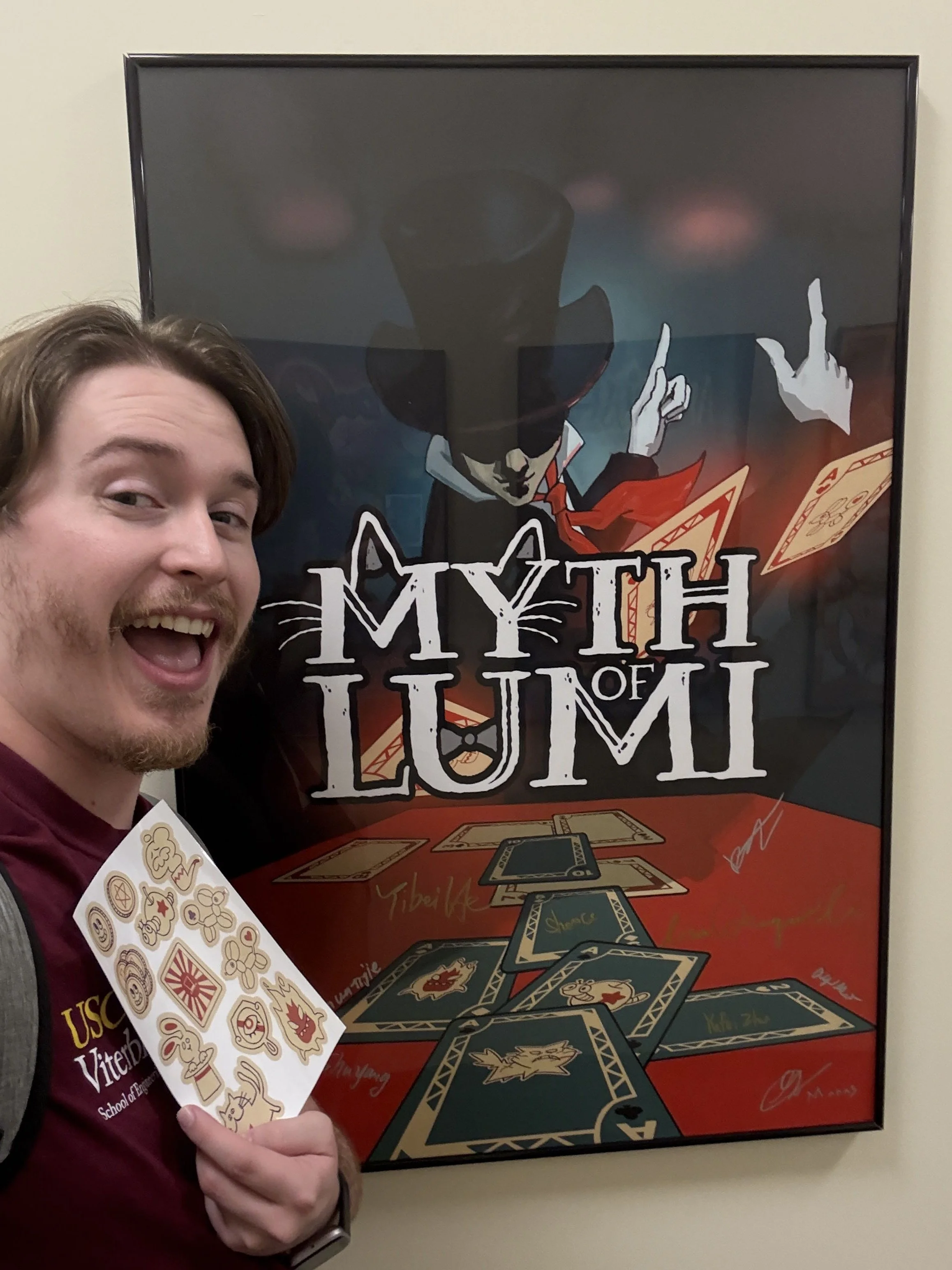 A young man smiling and holding a sheet of stickers with various designs. In the background, a poster titled 'Myth of Lumi' features a character with a top hat and playing cards.