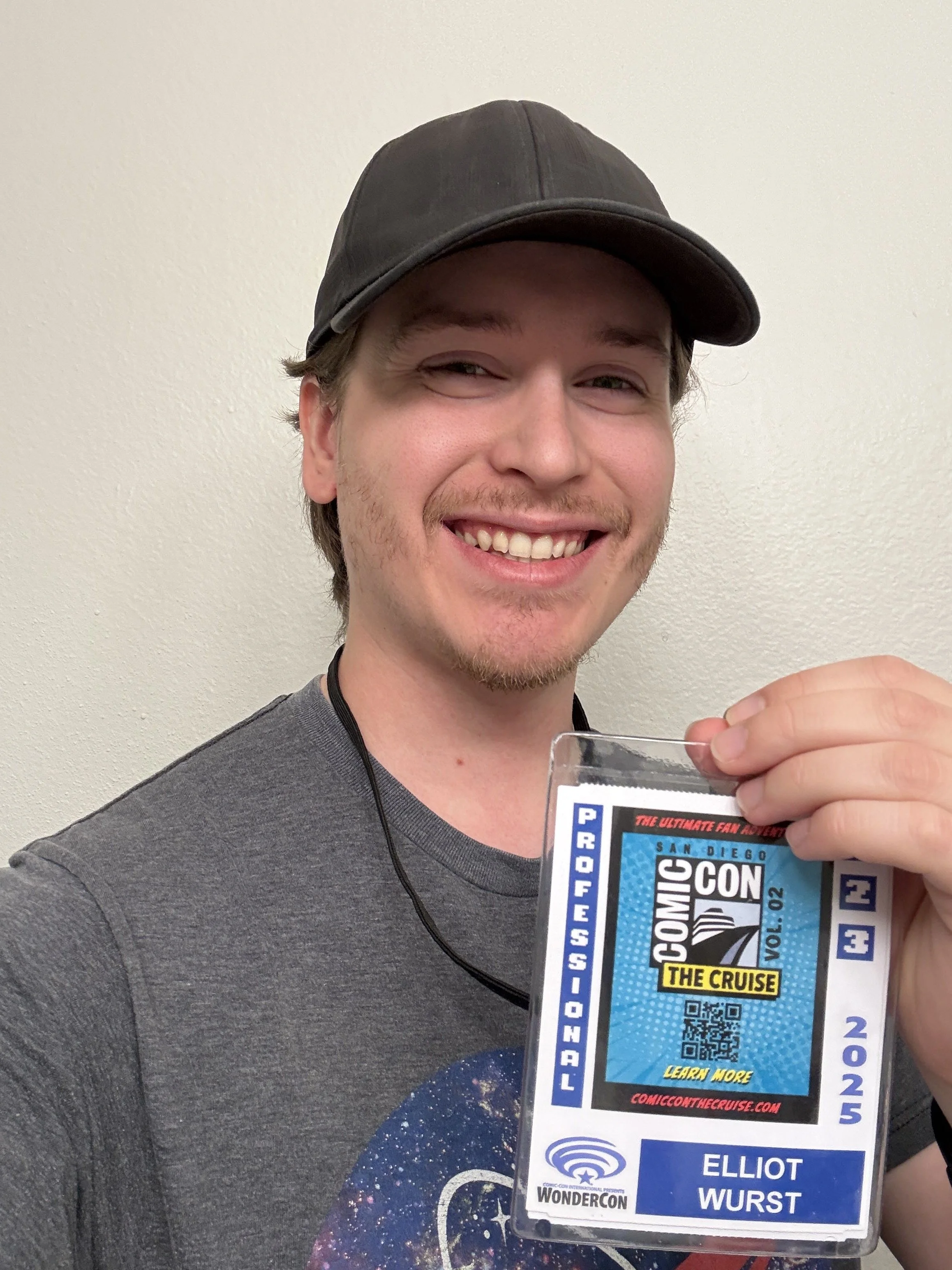A young man with a black cap and a gray T-shirt smiling and holding a badge for WonderCon 2025, featuring the cruise logo and his name Elliot Wurst.