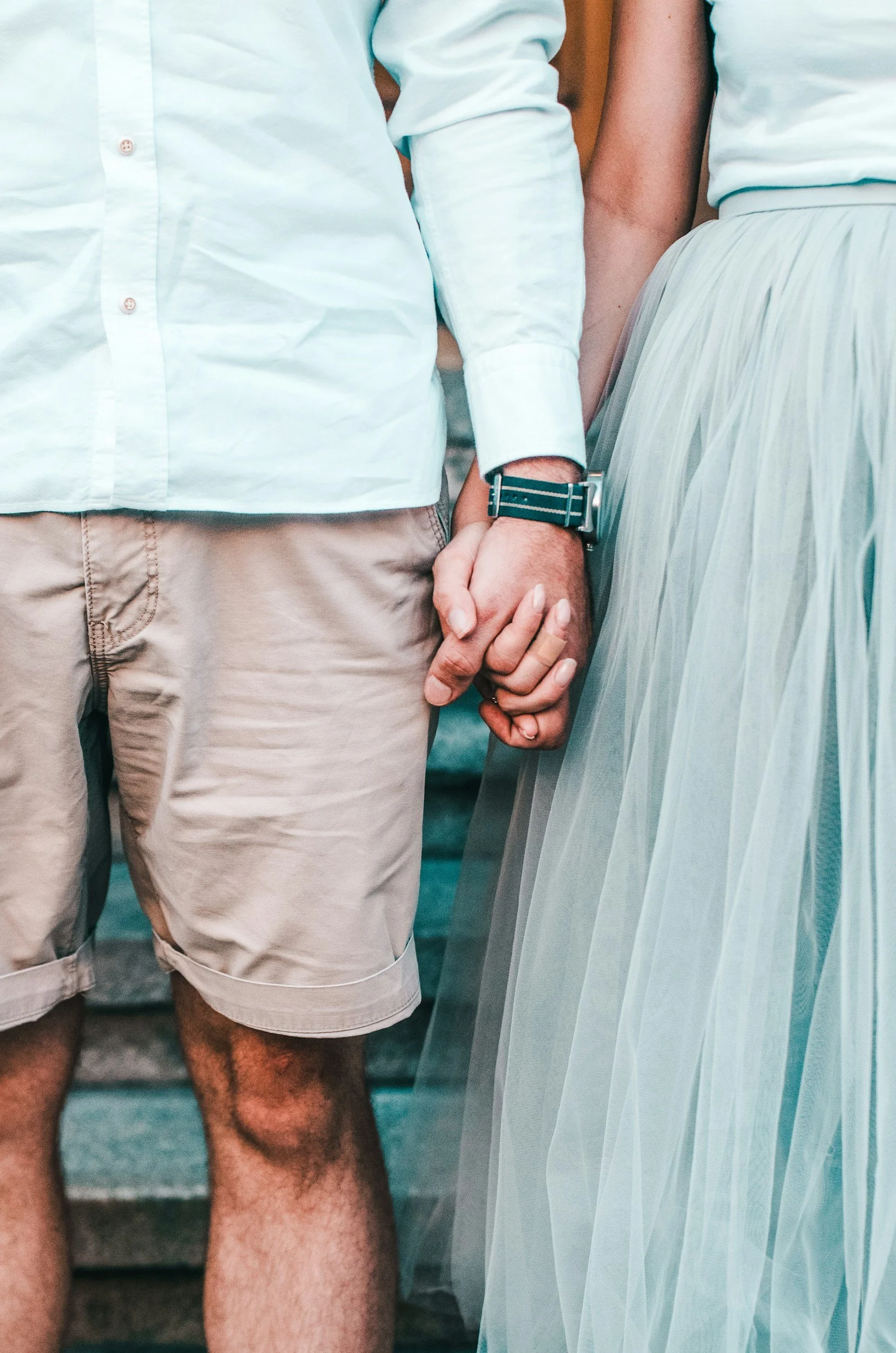 A close-up of a couple holding hands, with the man wearing beige shorts and a light blue shirt, and the woman wearing a light-colored skirt, on a set of stone stairs.