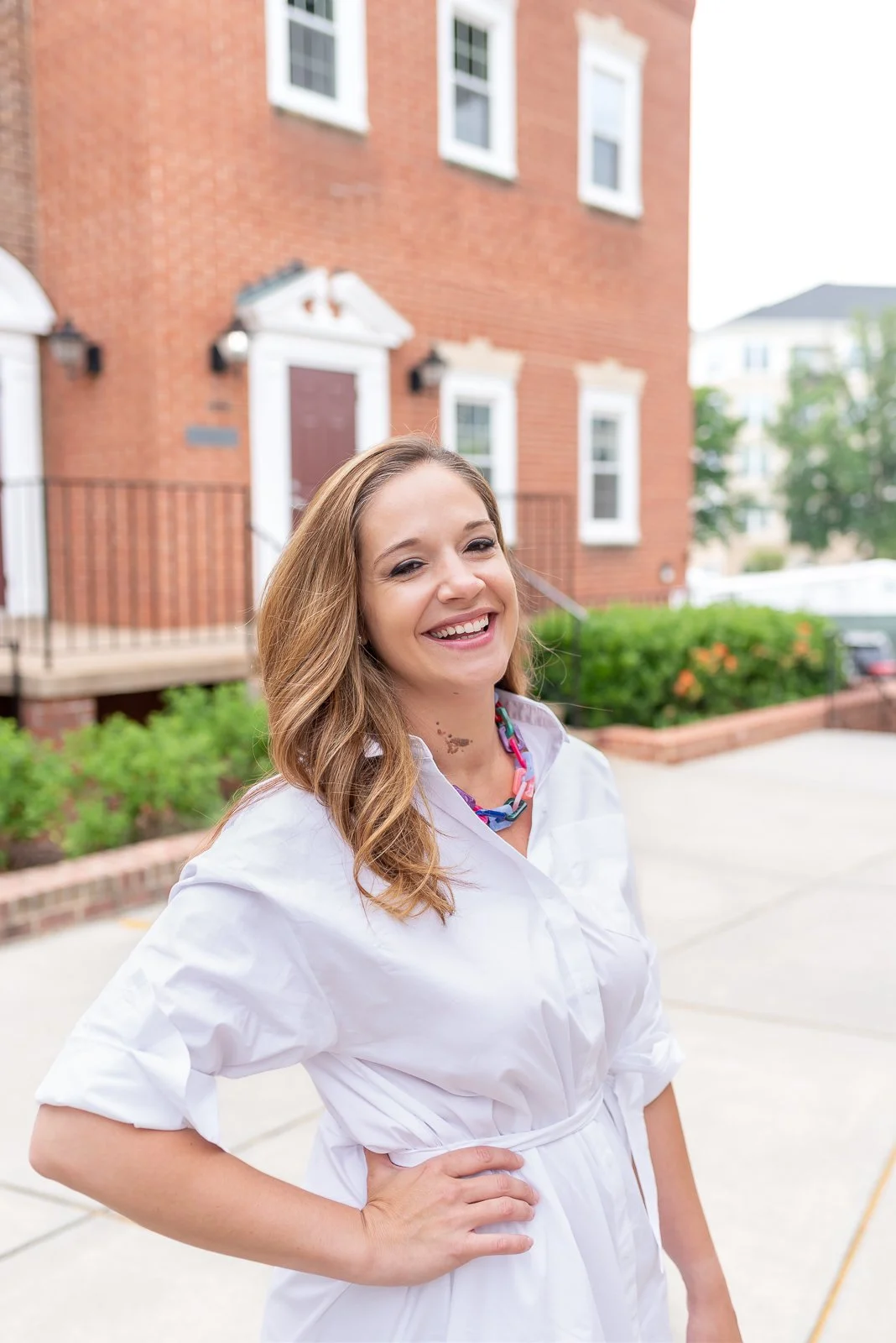 A young woman with long wavy hair, smiling, wearing white outfit and a colorful necklace, standing outdoors in front of a brick building with greenery.