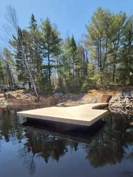 A wooden dock extends into a calm lake with clear blue water, surrounded by trees with green foliage, under a bright blue sky.