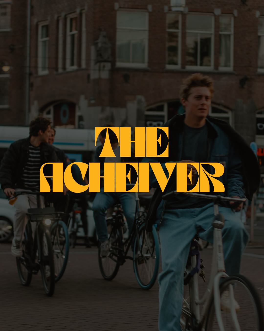 People riding bicycles on a city street at dusk with a brick building in the background and a yellow text overlay that reads 'The Achiever'.