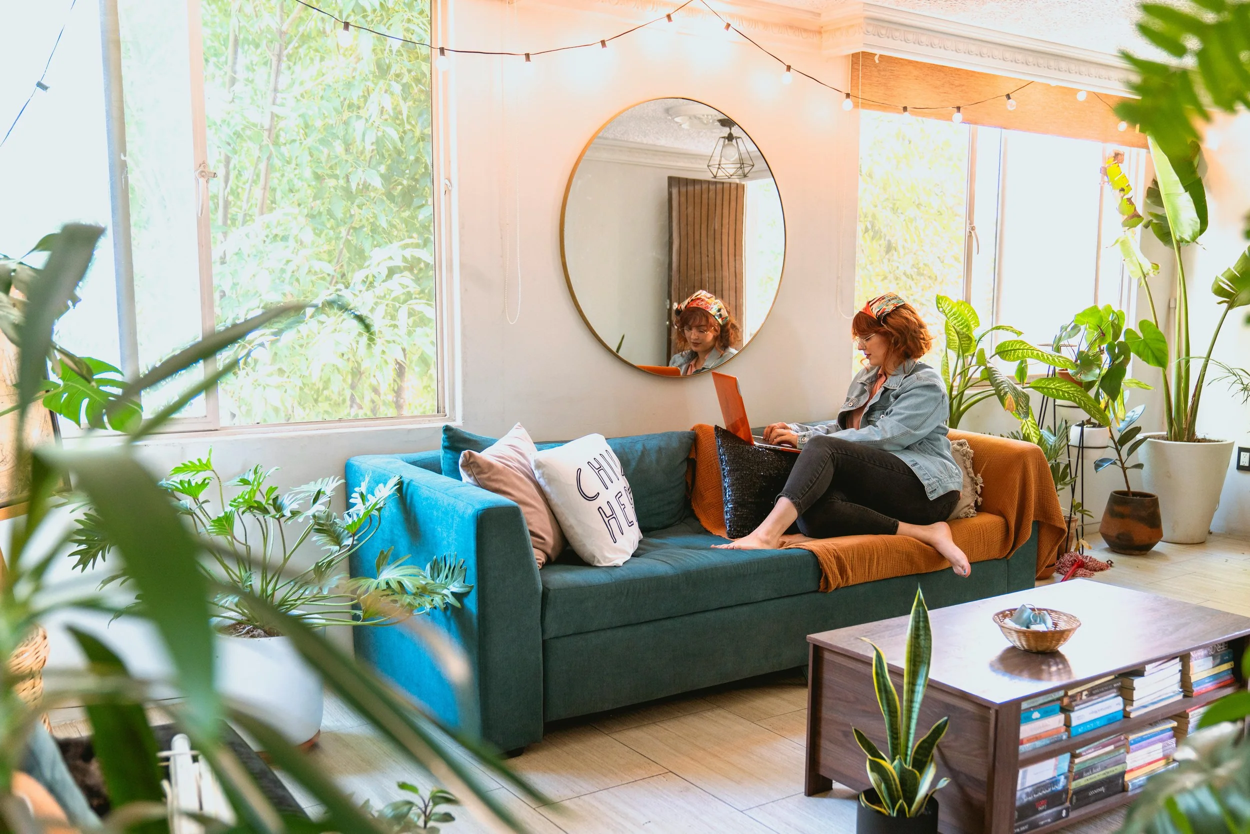 A woman with red hair sitting on a teal sofa in a living room, using a laptop, surrounded by plants and sunlight coming through large windows, with a round mirror on the wall behind her.