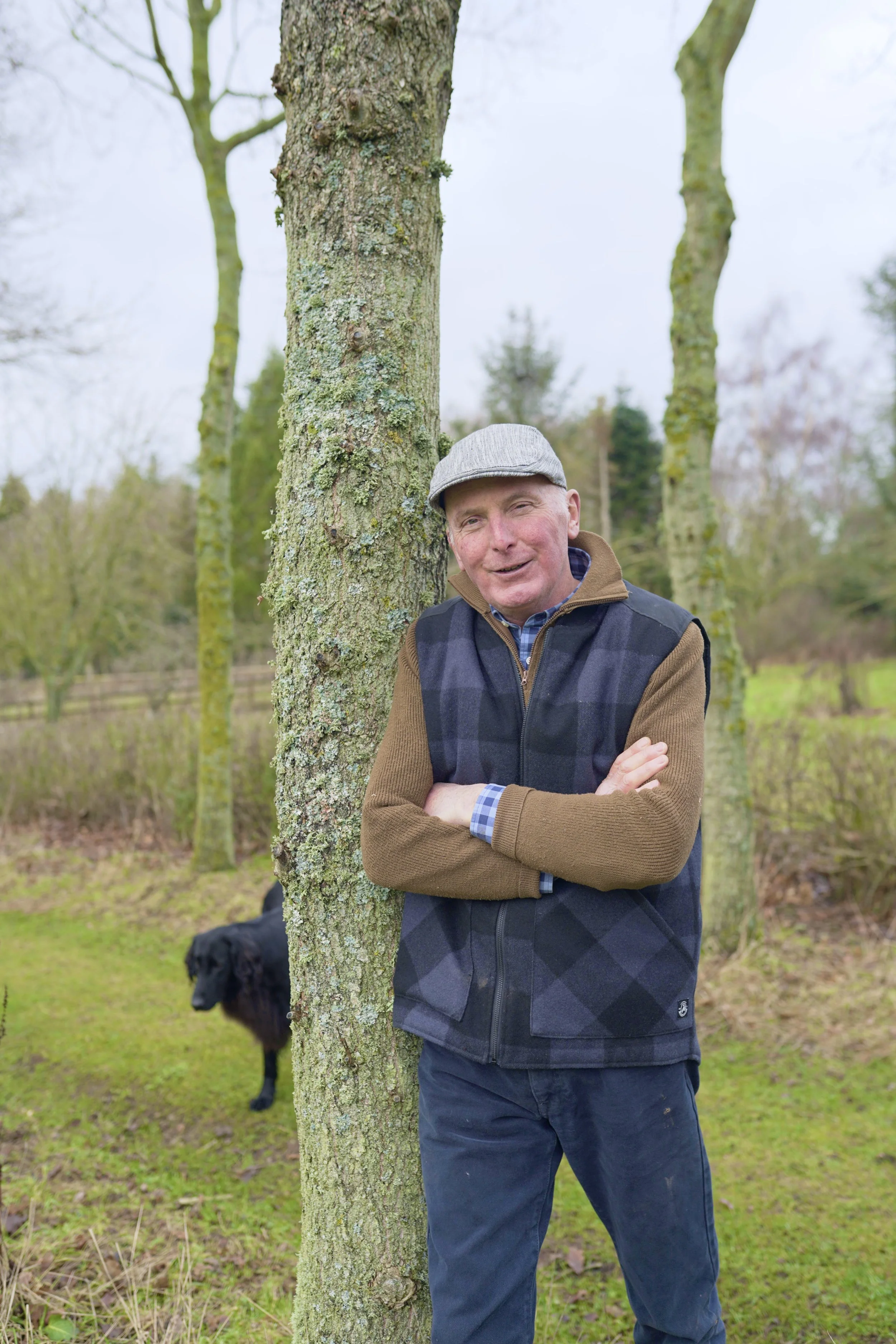 An elderly man leaning against a tree with a black dog in the background in a park or wooded area.