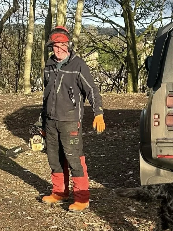 A man dressed in outdoor work gear, holding a chainsaw, standing on a wooded dirt path next to a parked car, with a black dog nearby.