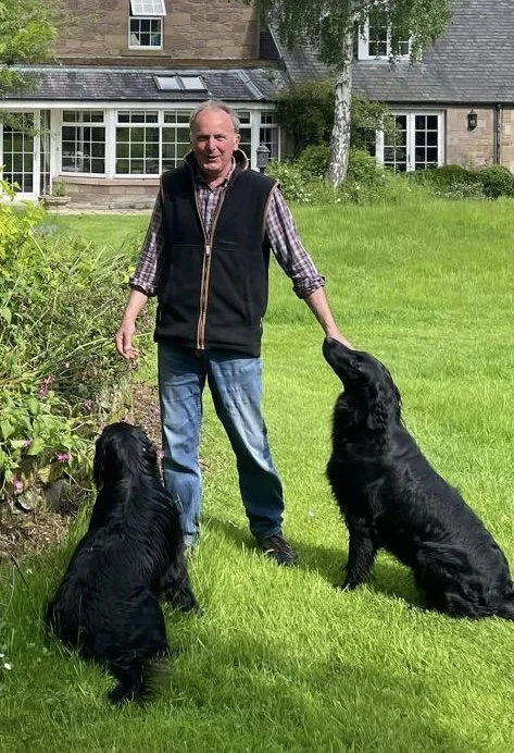An elderly man standing on a lush green lawn with two large black retrievers, one sitting and one standing, near a garden bed with plants and flowers, in front of a large house with multiple windows and a porch.