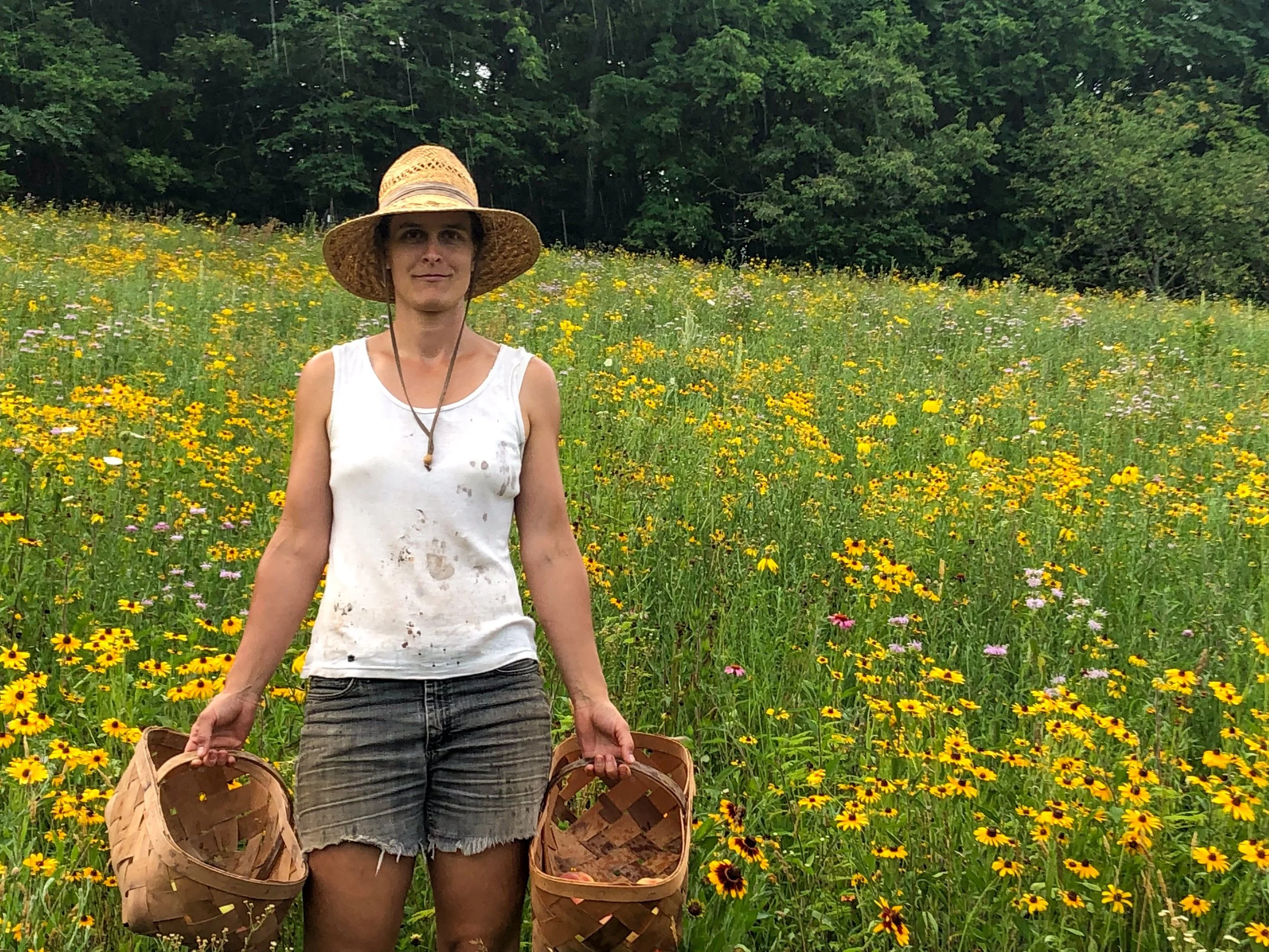 A woman standing in a field of mostly yellow wildflowers, wearing a wide-brimmed straw hat, a white sleeveless top, and black shorts, holding two woven baskets.