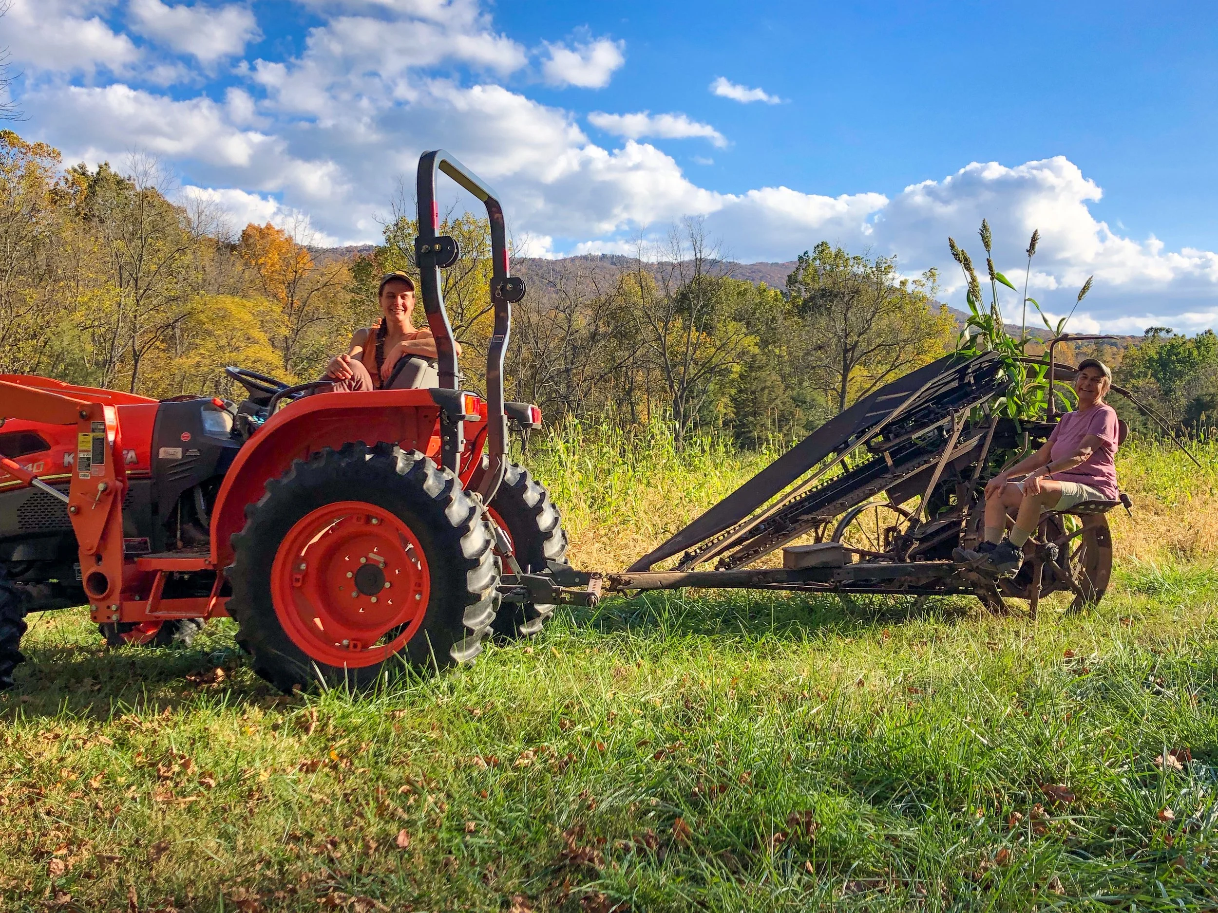 Two people sit on a farm tractor and a piece of farming equipment with a backdrop of trees and mountains on a partly cloudy day.