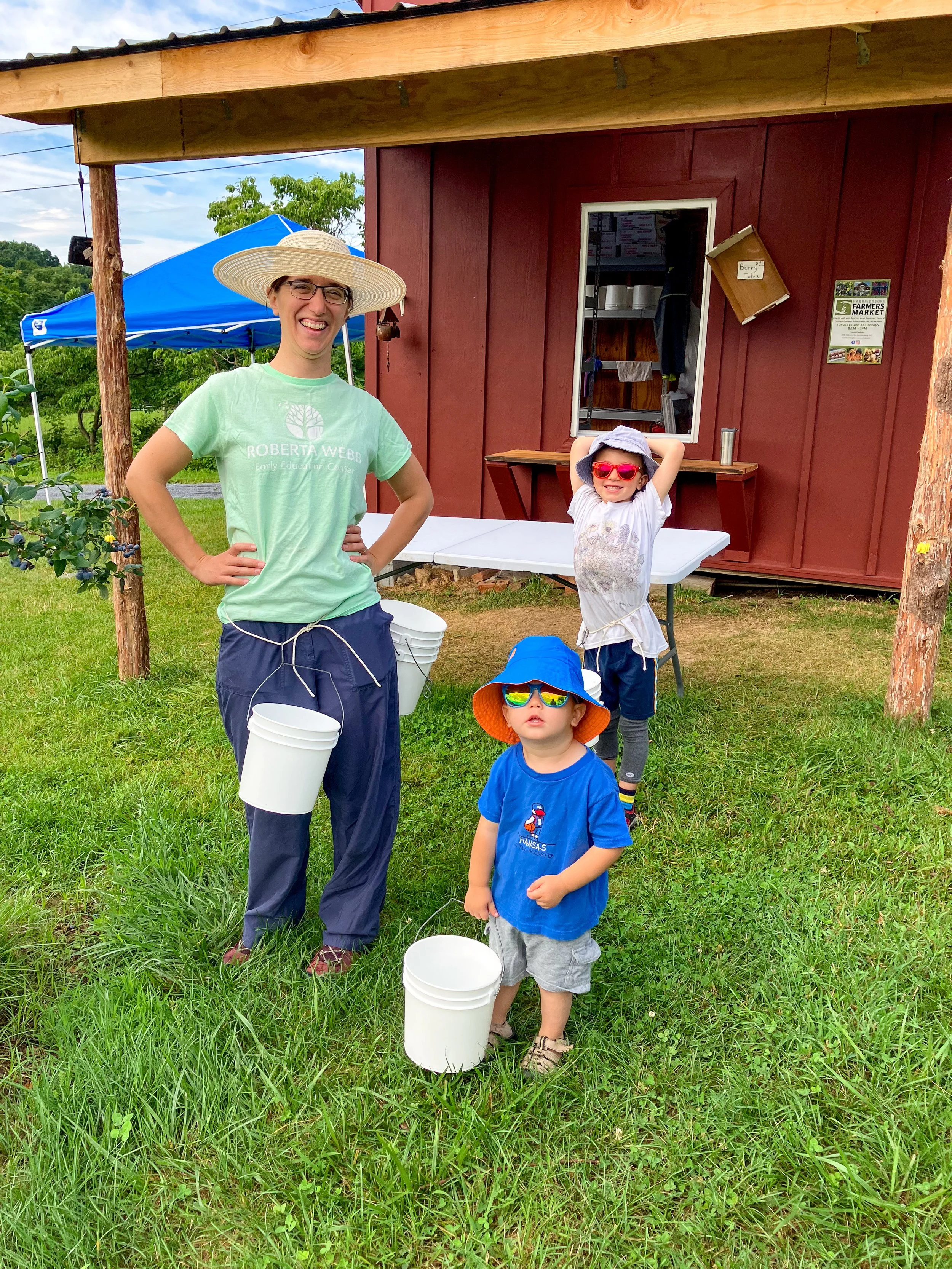 A woman and two young children standing on grass outdoors in front of a small farm stand, with buckets and wearing sun hats and sunglasses.