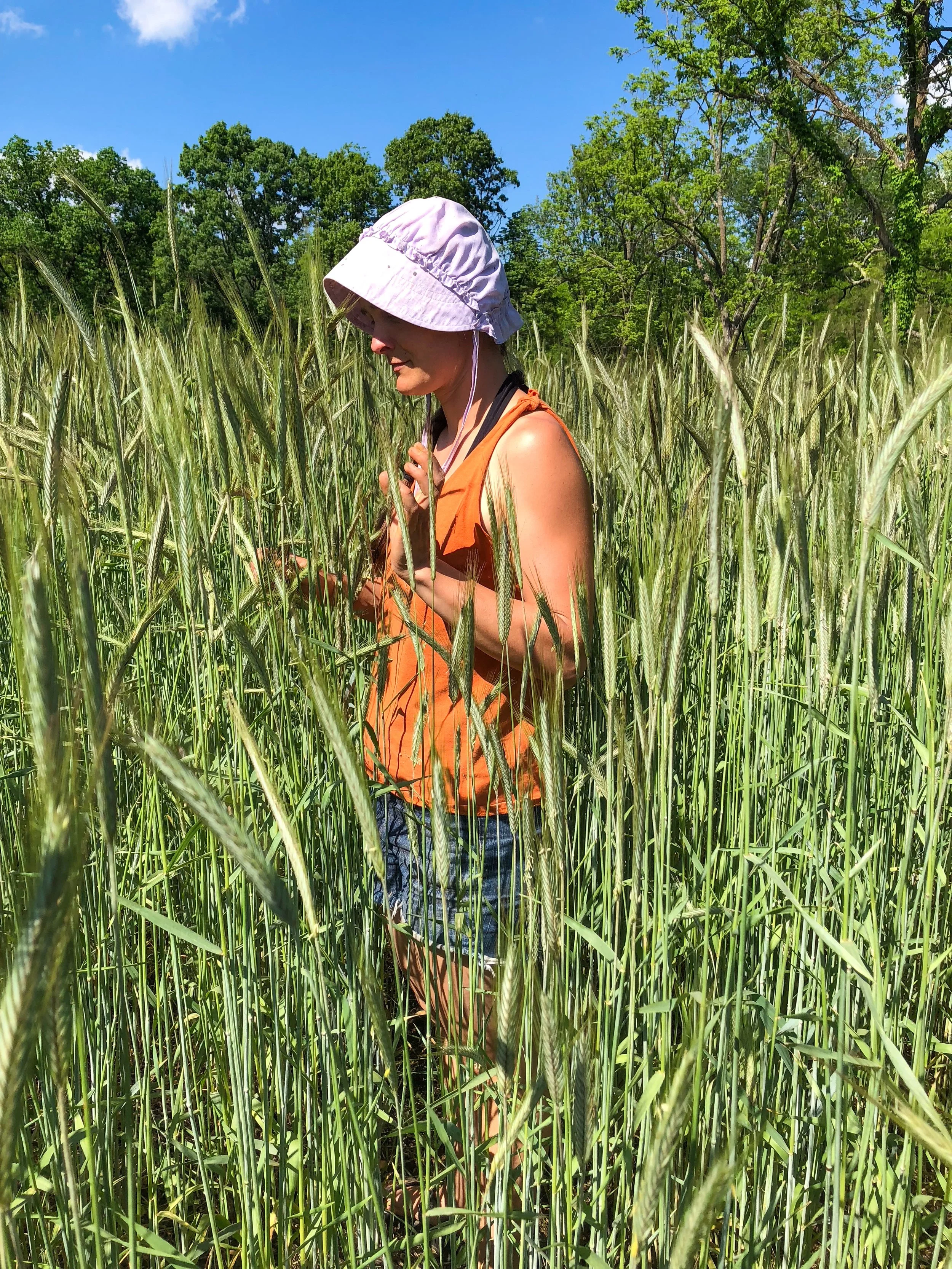 A woman in an orange tank top, denim shorts, and a light-colored sun hat walking through a green field on a sunny day.