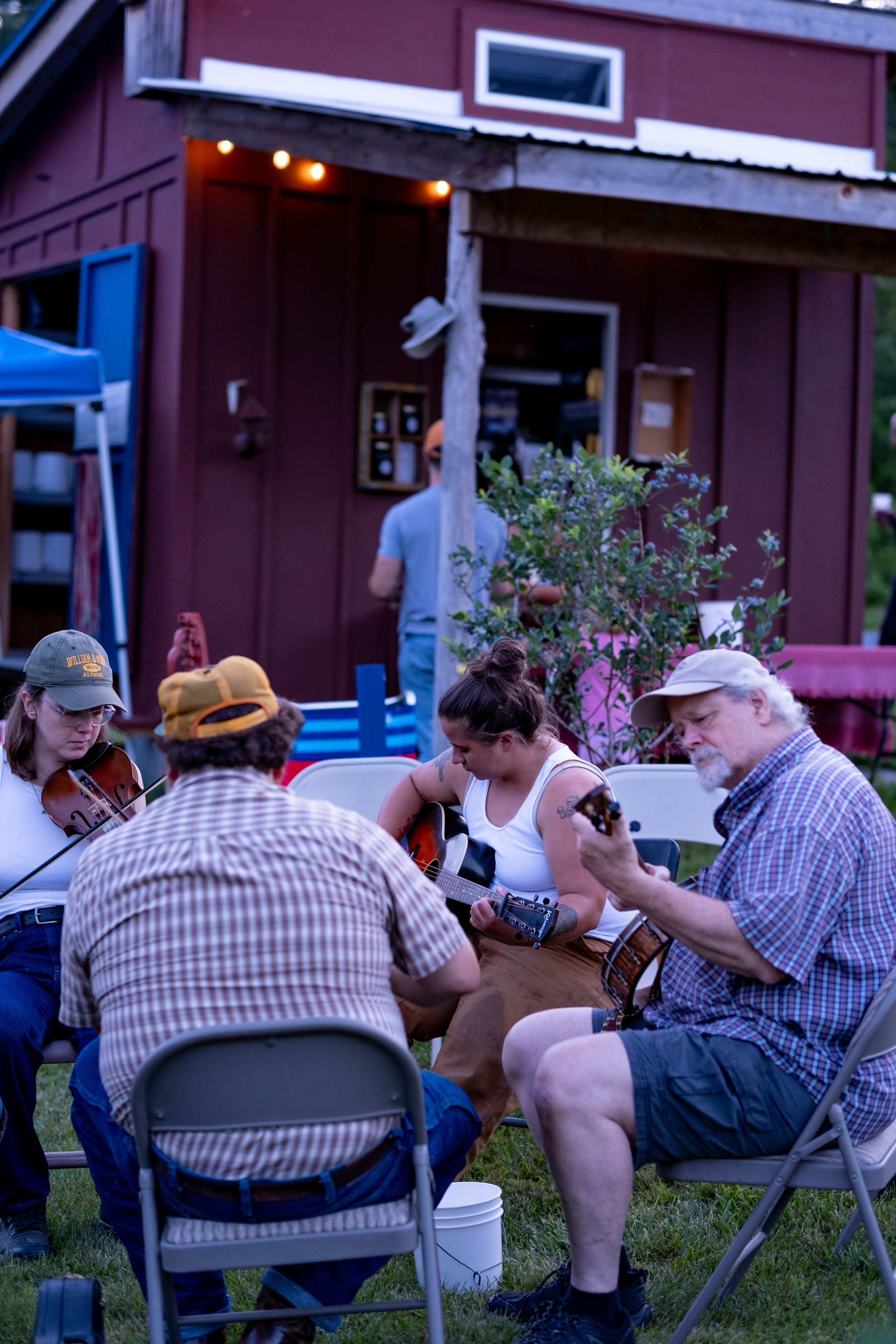 A group of people playing musical instruments at an outdoor gathering in front of a red farm building with string lights.