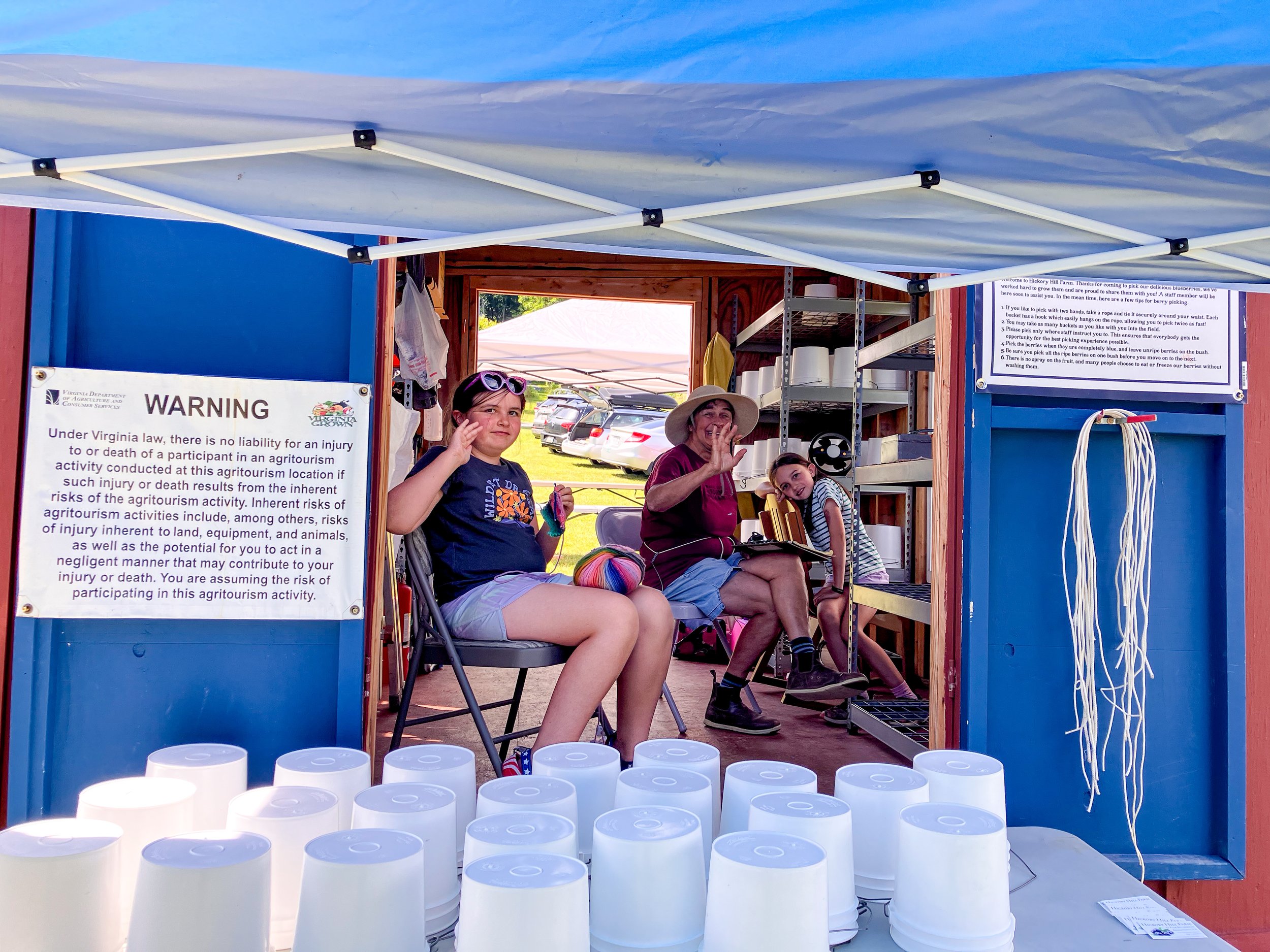 Three people sitting inside a blue wooden farm building, with one person waving and another looking at the camera. There are cars parked outdoors in the background.
