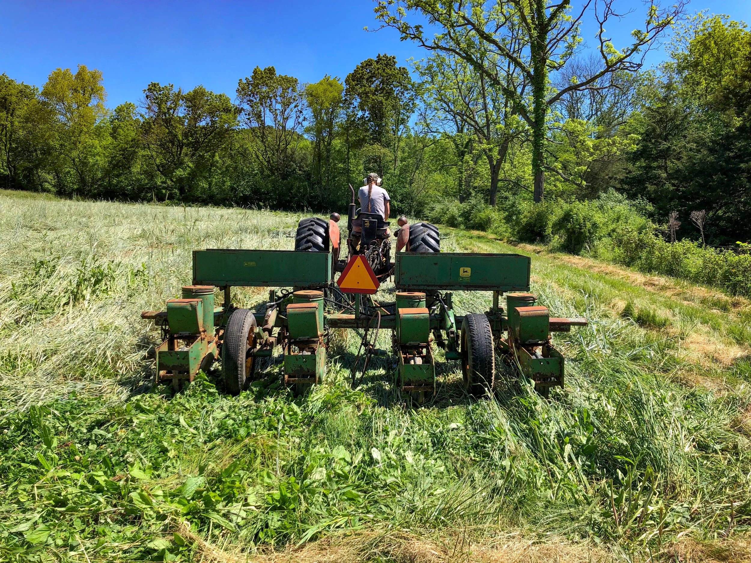 A person driving a green tractor with attached equipment in a grassy field surrounded by trees under a clear blue sky.