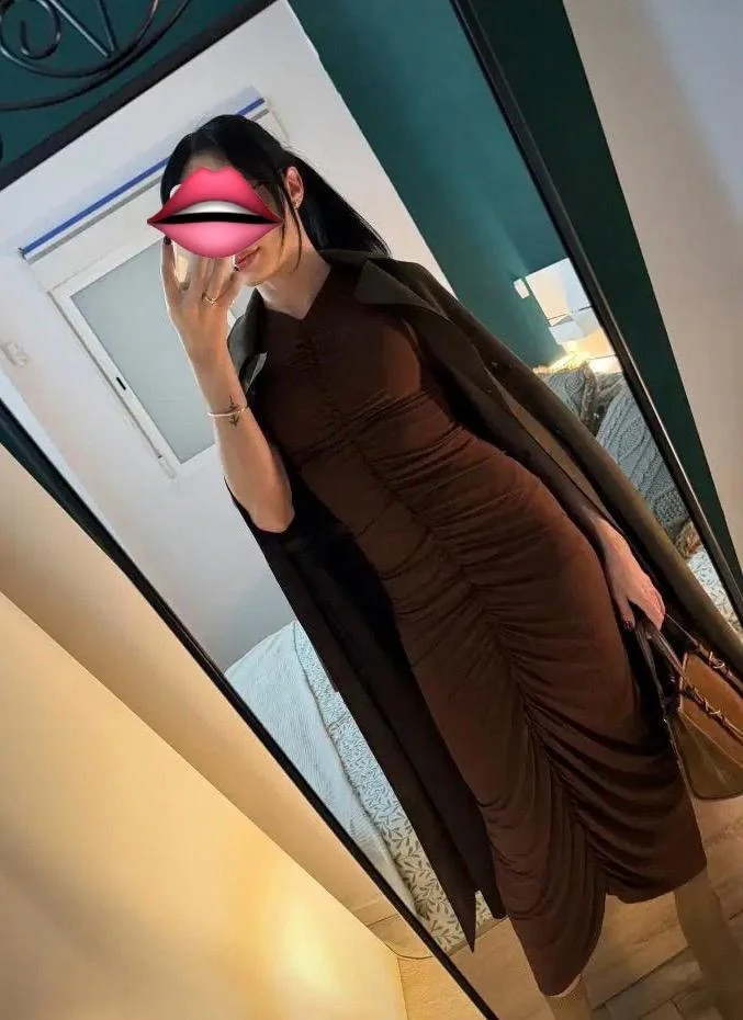 A woman standing outdoors at night, wearing a white fringed dress, with her back to the camera. She has long dark hair and is smiling, with city lights in the background. Tantra Elite Escort Amsterdam 