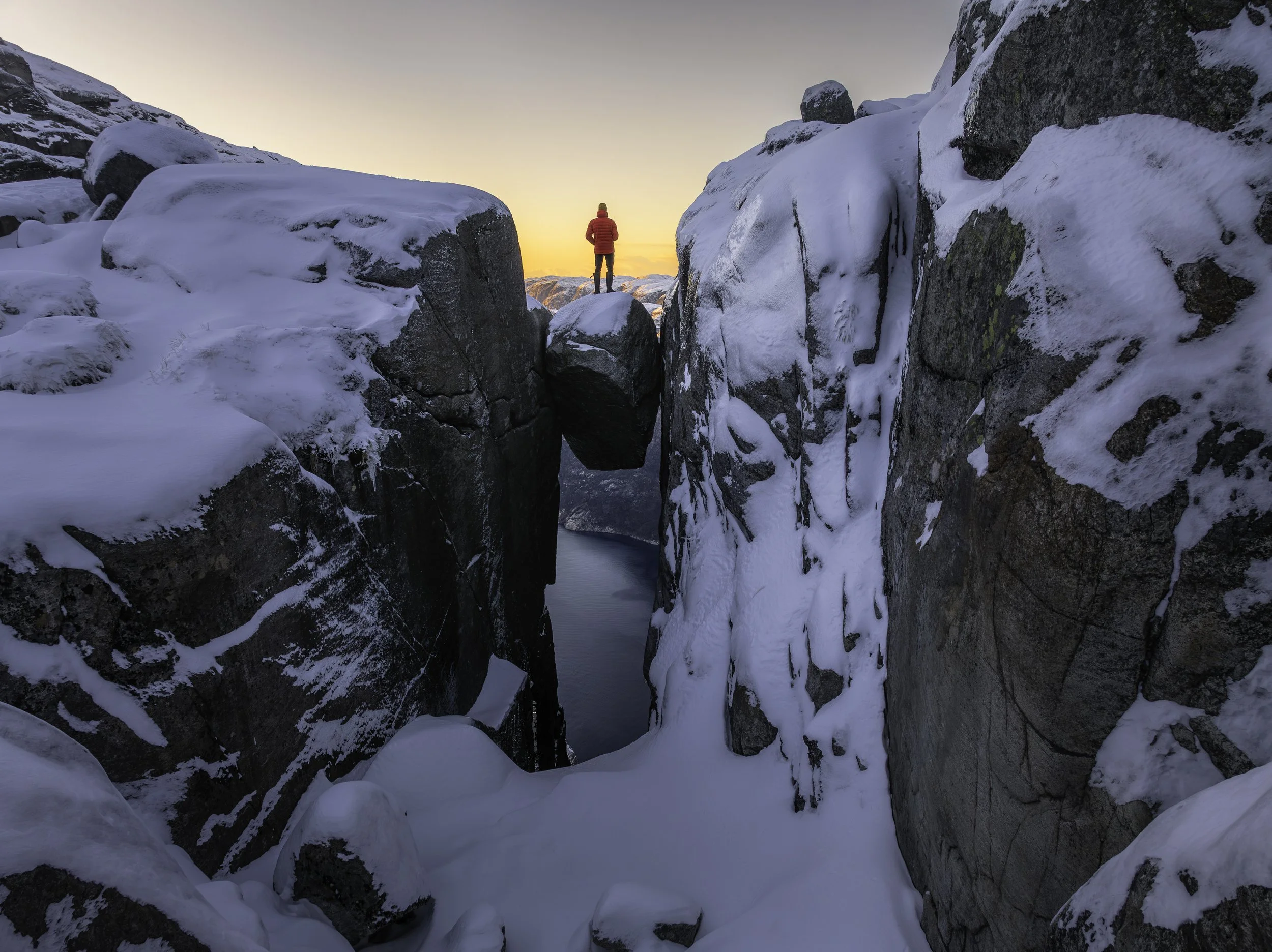 A person in a red jacket standing on a snow-covered rock between tall icy cliffs at sunset or sunrise.