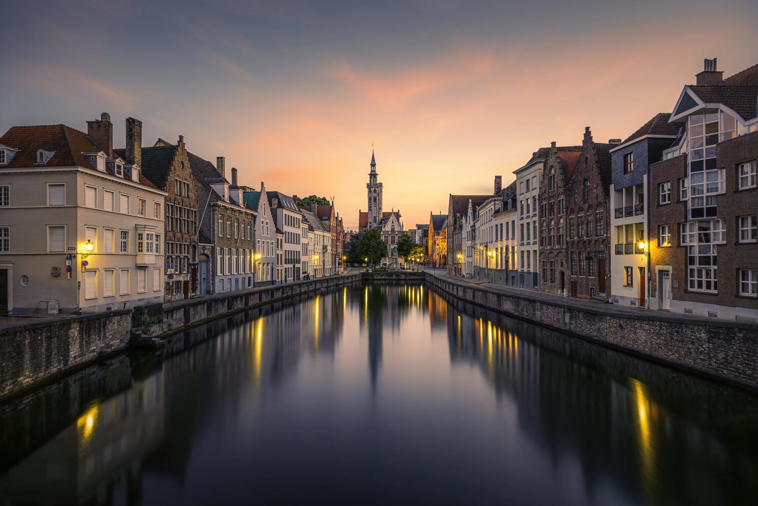 A canal with historic European-style buildings on either side, reflective water, soft sunset sky, and a church tower in the background.