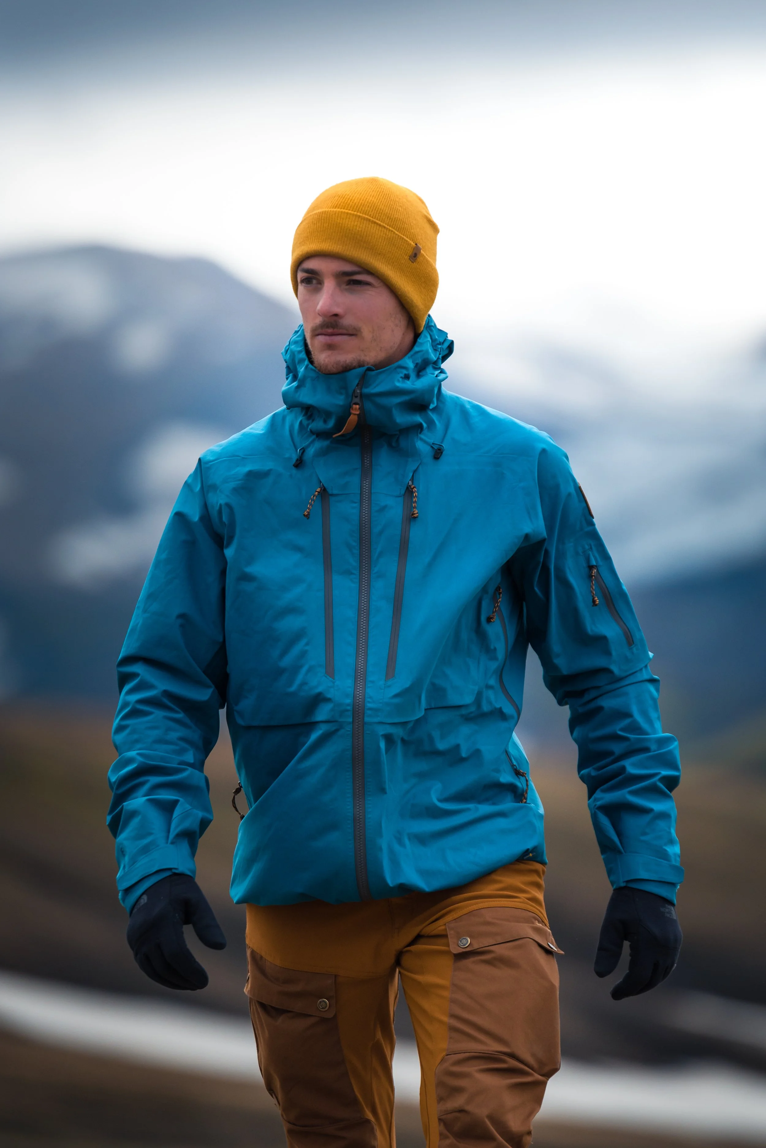 A man dressed in a blue insulated jacket, yellow beanie, and brown pants stands outdoors in a mountainous landscape under an overcast sky.