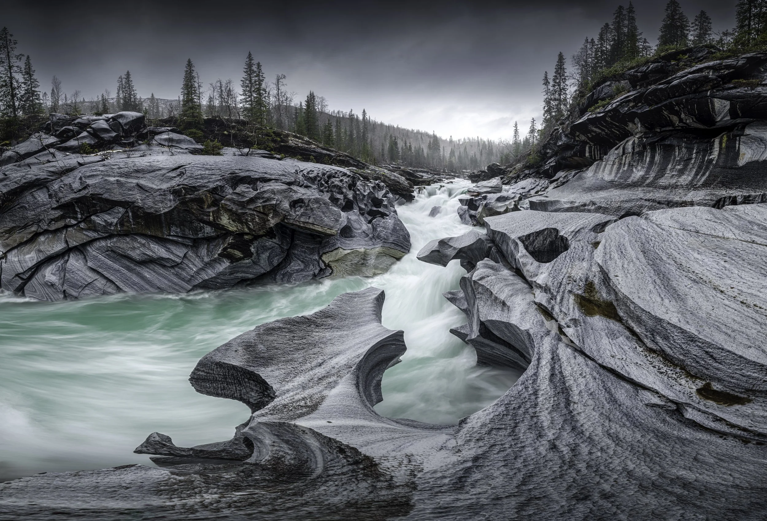 Flowing river through rocky canyon with pine trees and a cloudy sky.