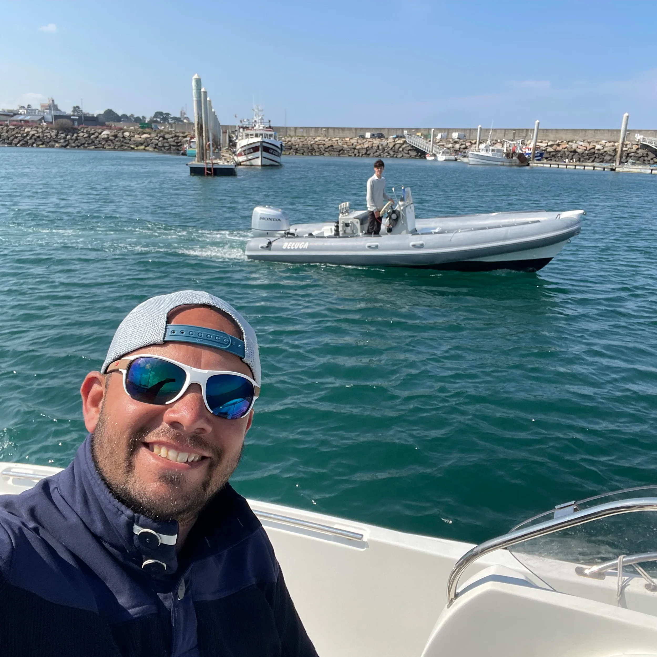 Un homme souriant portant des lunettes de soleil et une casquette sur un bateau à moteur, avec une mer calme et un autre bateau en arrière-plan.
