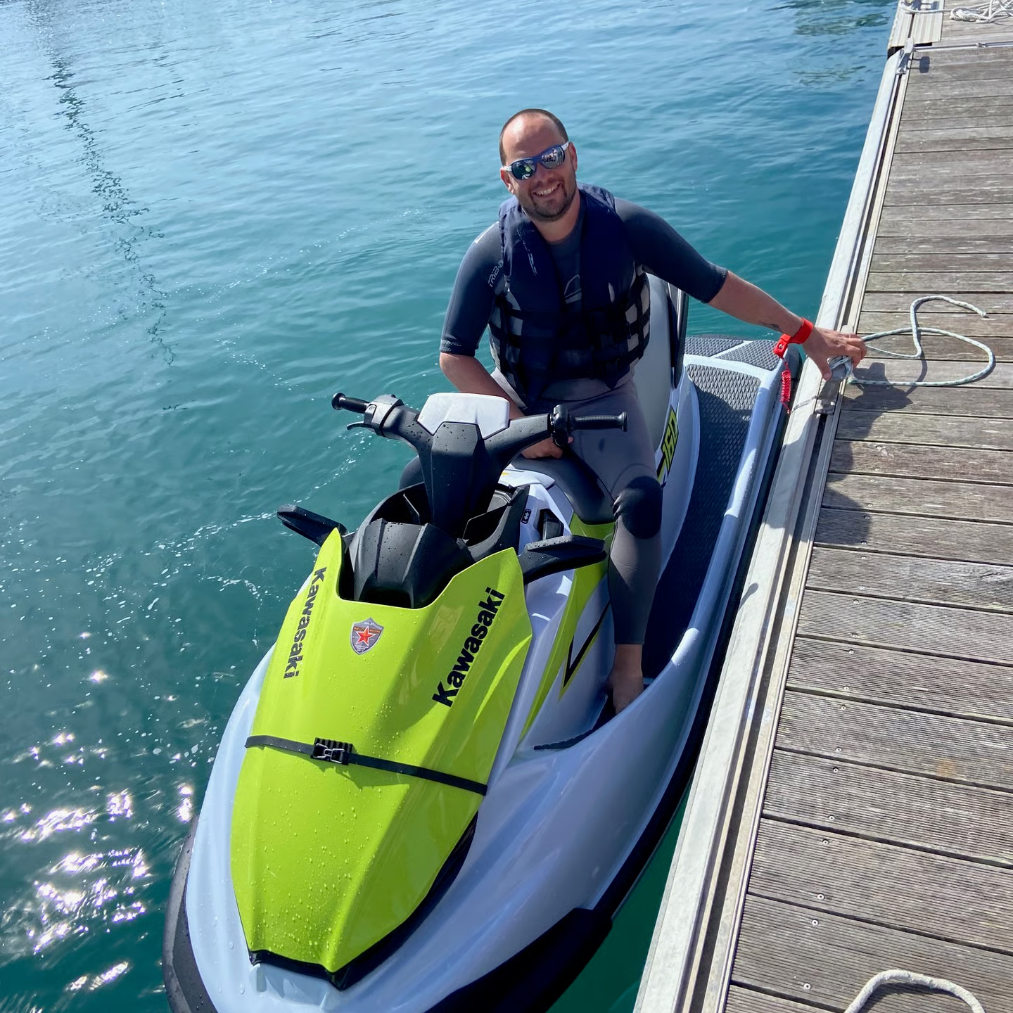 Un homme souriant portant des lunettes de soleil, assis sur une moto-neige verte et blanche attachée à un quai en bois, près d'une étendue d'eau calme.
