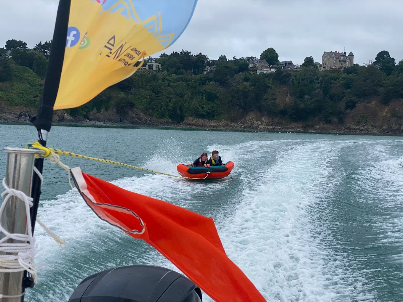 Deux personnes sur une bouée motorisée sur une mer calme avec des maisons en hauteur dans la forêt en arrière-plan.