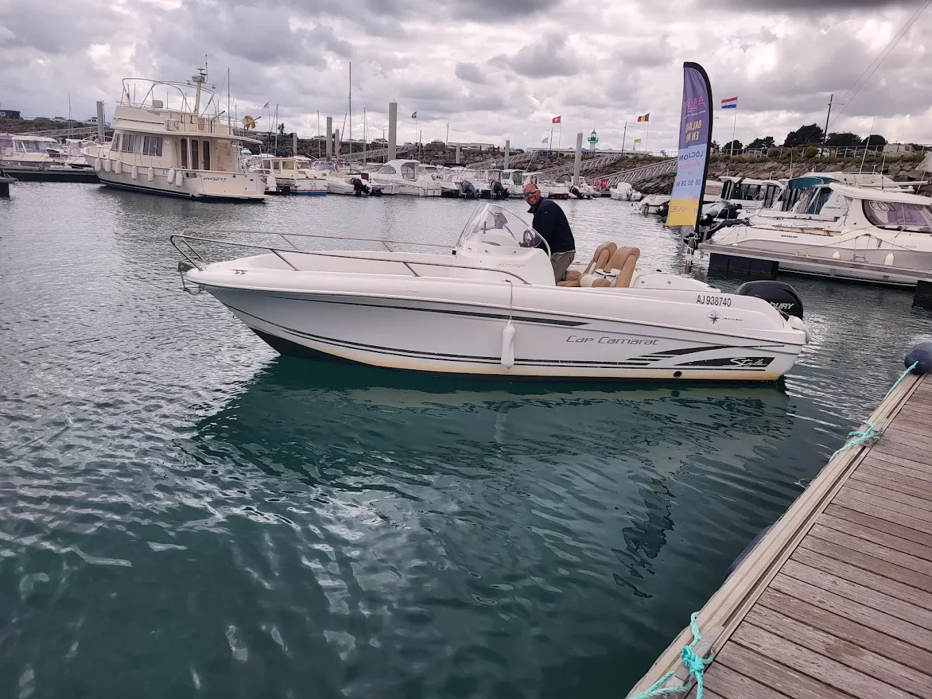 Une personne navigue en bateau à moteur dans un port avec plusieurs yachts et voiliers amarrés. Le ciel est nuageux.