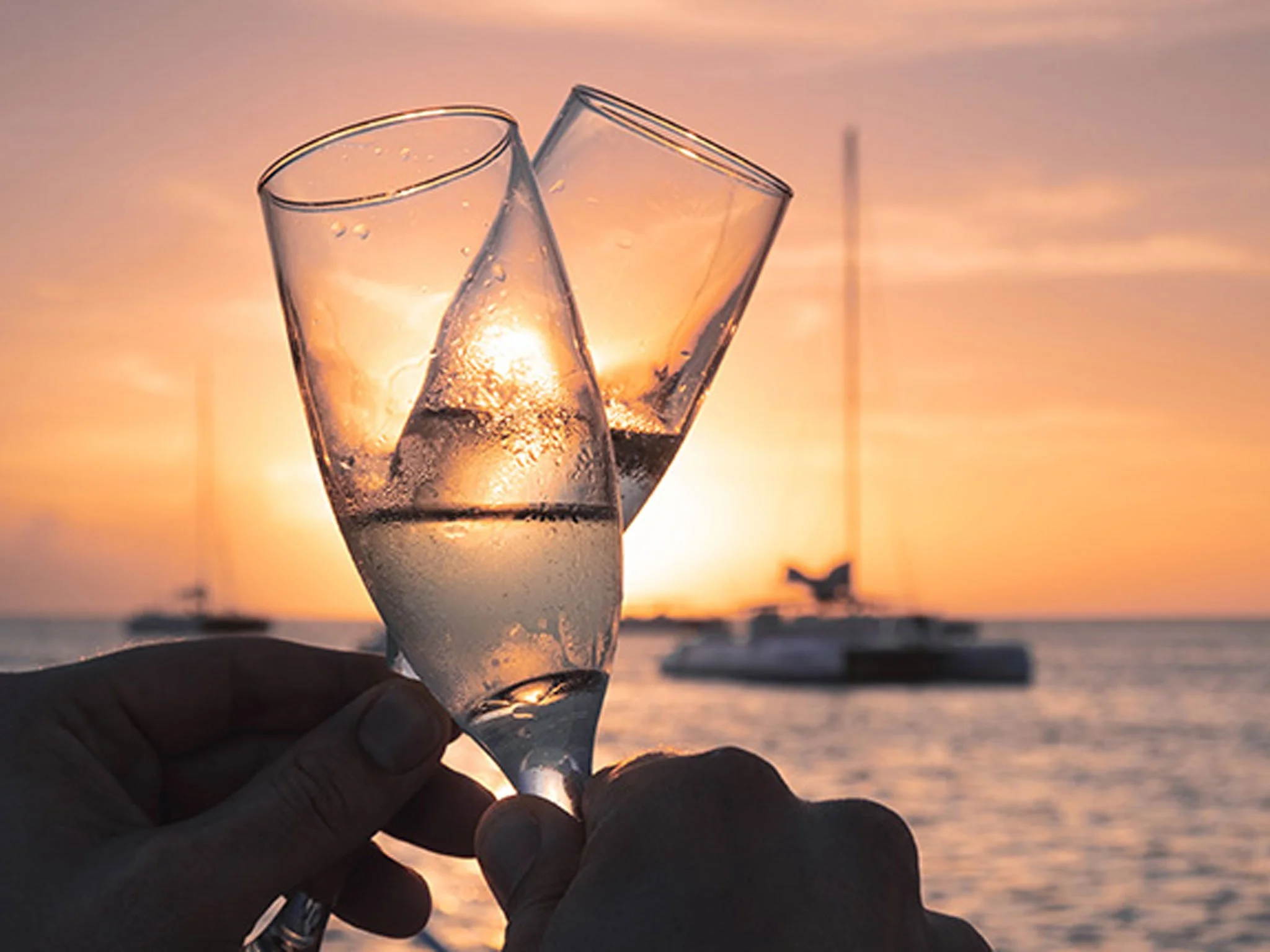 Verres de champagne portés par deux personnes lors d'un coucher de soleil sur la mer, avec un bateau en arrière-plan.