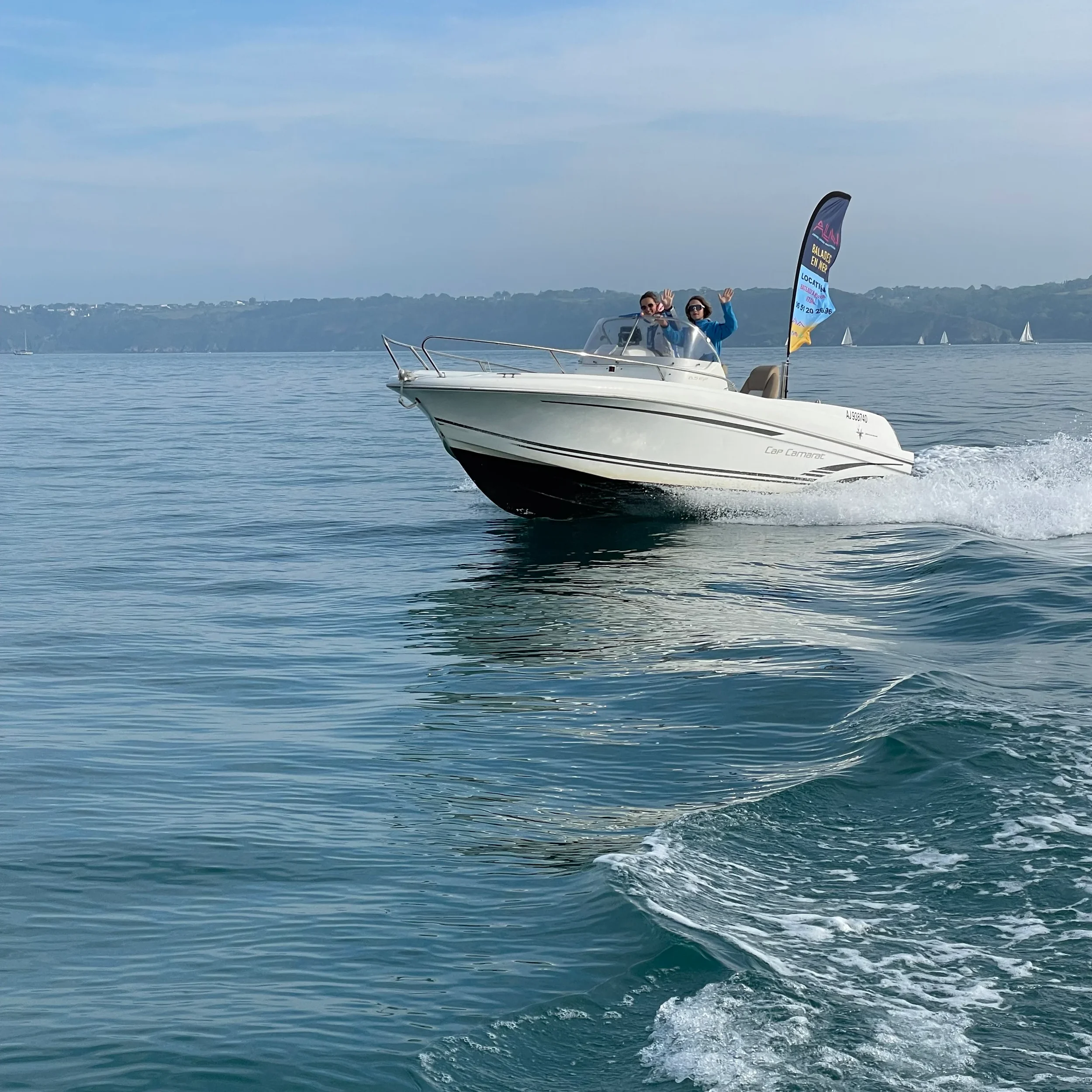 Une bateau à moteur avec deux personnes à bord navigue sur l'eau calme, avec un drapeau publicitaire flottant à l'arrière.
