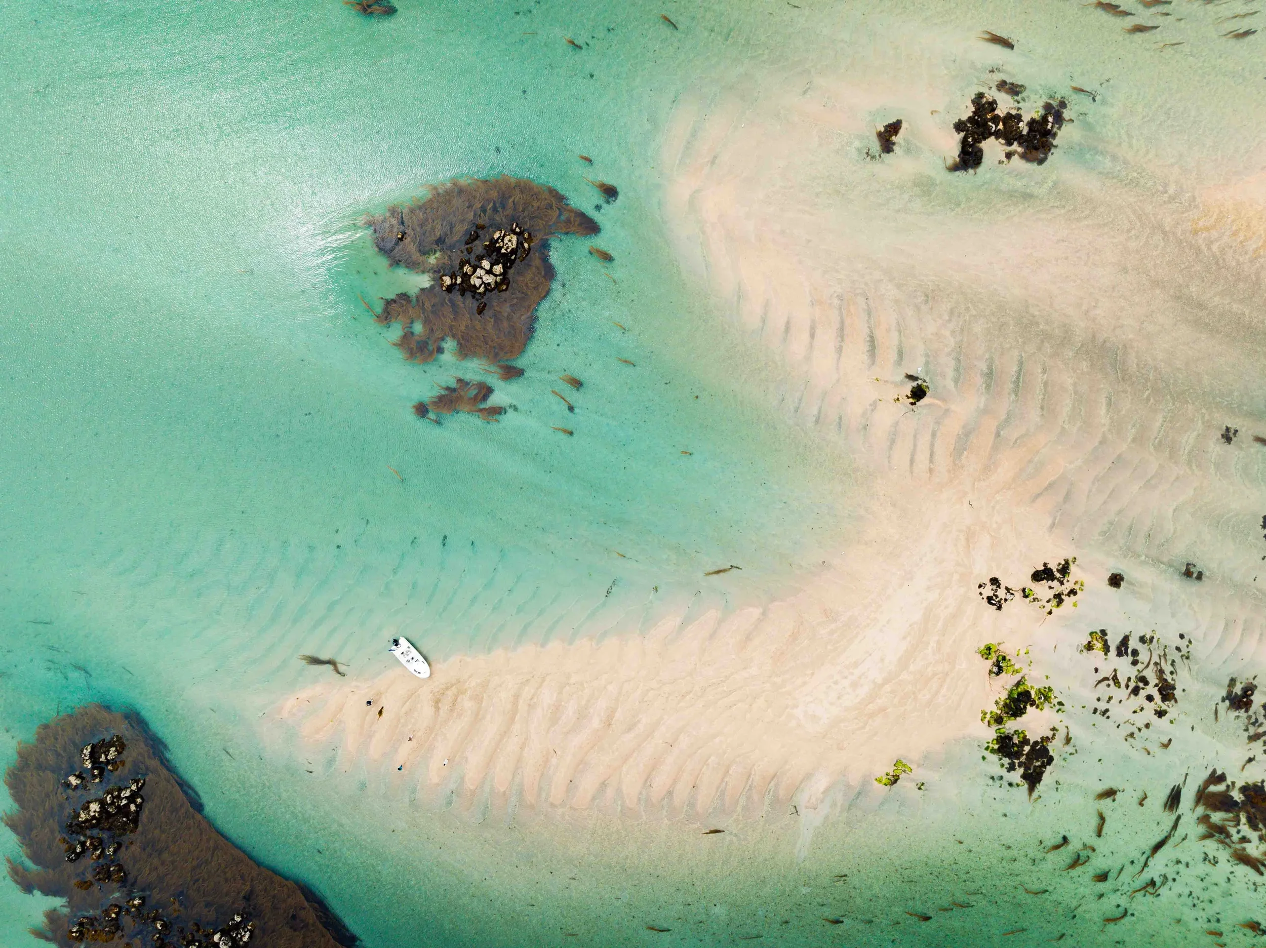 Vue aérienne d'une plage de sable blanc avec des rochers soumis à la marée, eau turquoise, et un bateau sur la plage.