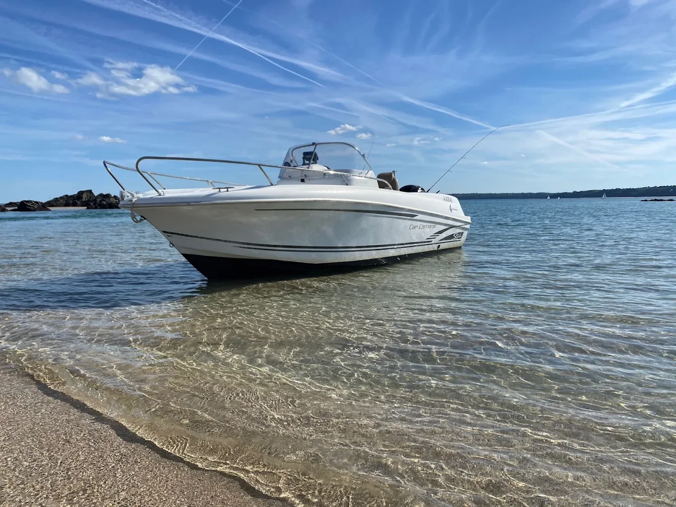 Bateau à moteur blanc ancré dans une eau calme à la plage sous un ciel bleu avec quelques nuages et traînées de condensation.
