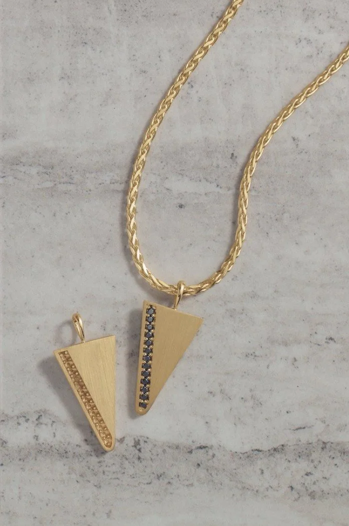 Gold necklace with two geometric pendants, one with embedded black stones, on a light marble surface.