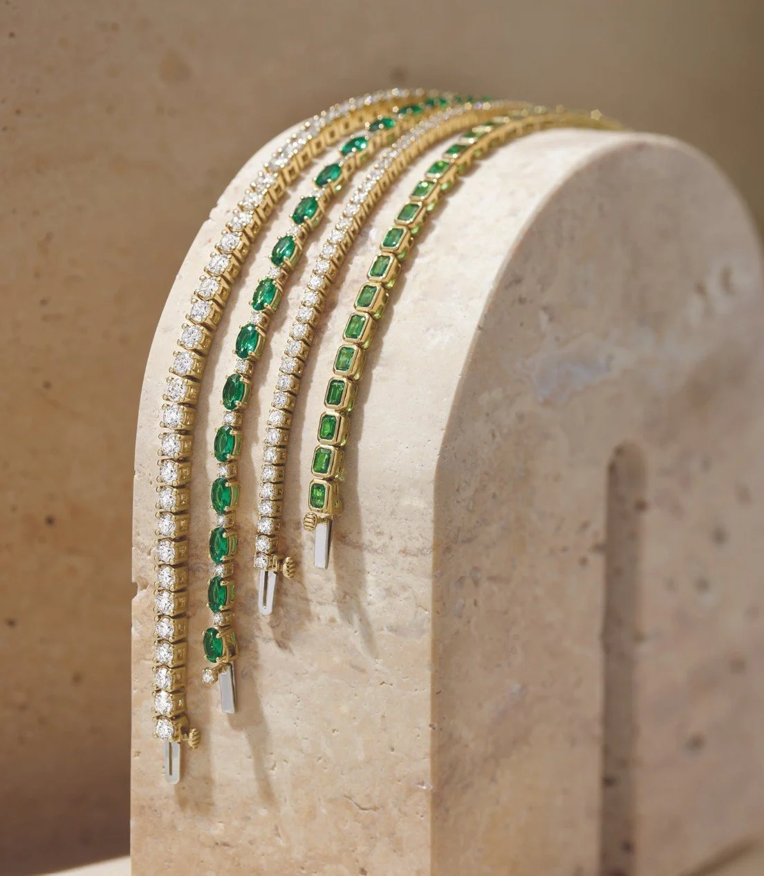 Four gold necklaces with green gemstones and clear diamonds displayed on a beige stone stand.