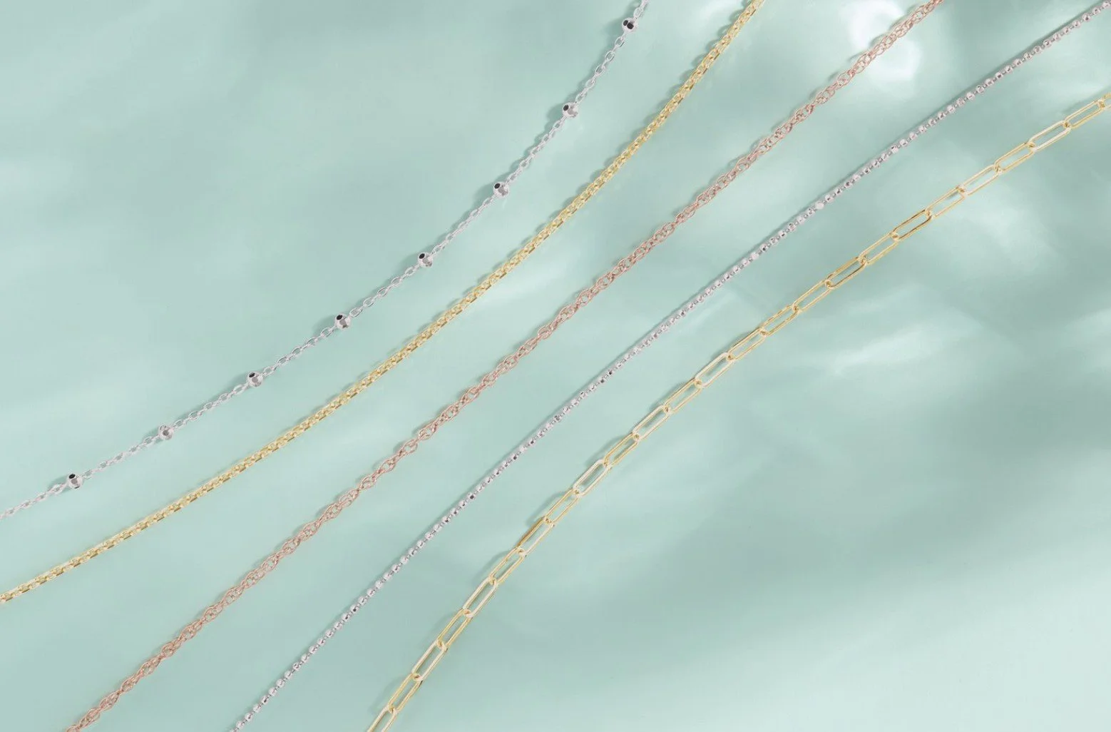 Multiple delicate chain necklaces in silver, gold, rose gold, and mixed metals laid out on a light blue surface.