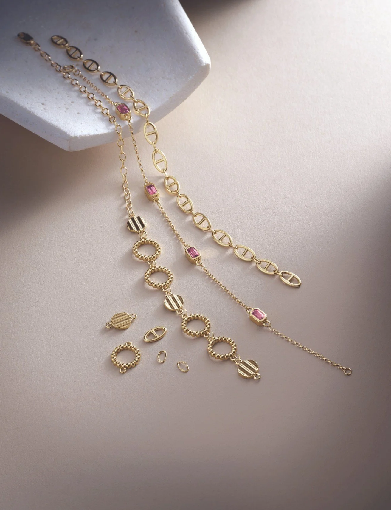 Three gold chains with various links and pink gemstones, and gold charm pieces, laid out on a light beige surface with a white textured box.