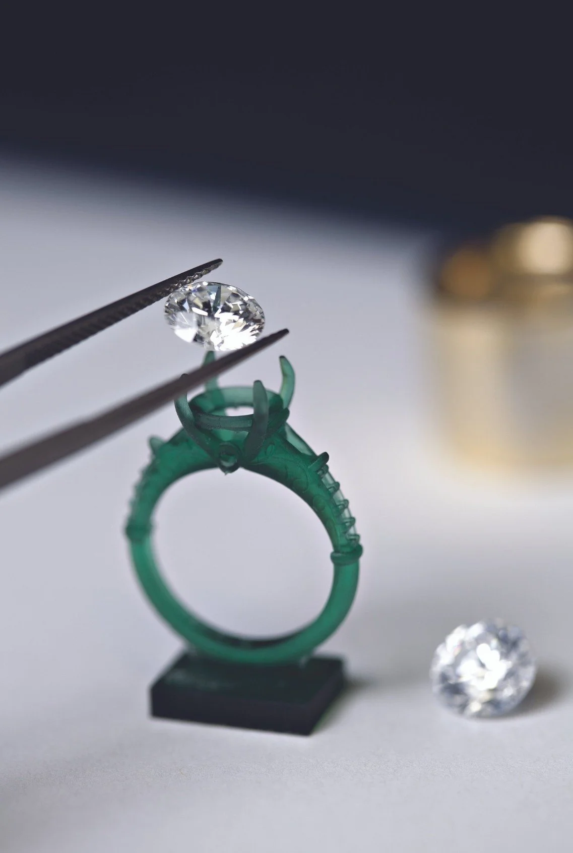 Jewelry setting with a diamond being placed by tweezers, a green ring in the background, and another diamond on the surface.
