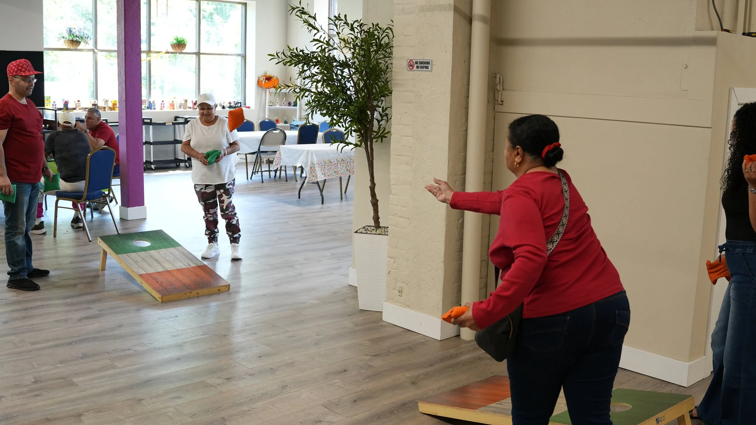 People playing cornhole in a brightly lit indoor space with tables and large windows in the background.
