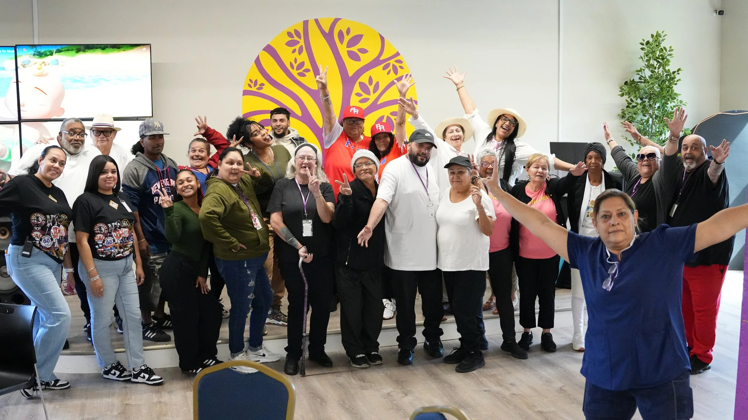 A diverse group of people gathered indoors, smiling and posing for a group photo with some making peace signs and others raising their arms in celebration, against a yellow and purple decorative backdrop.