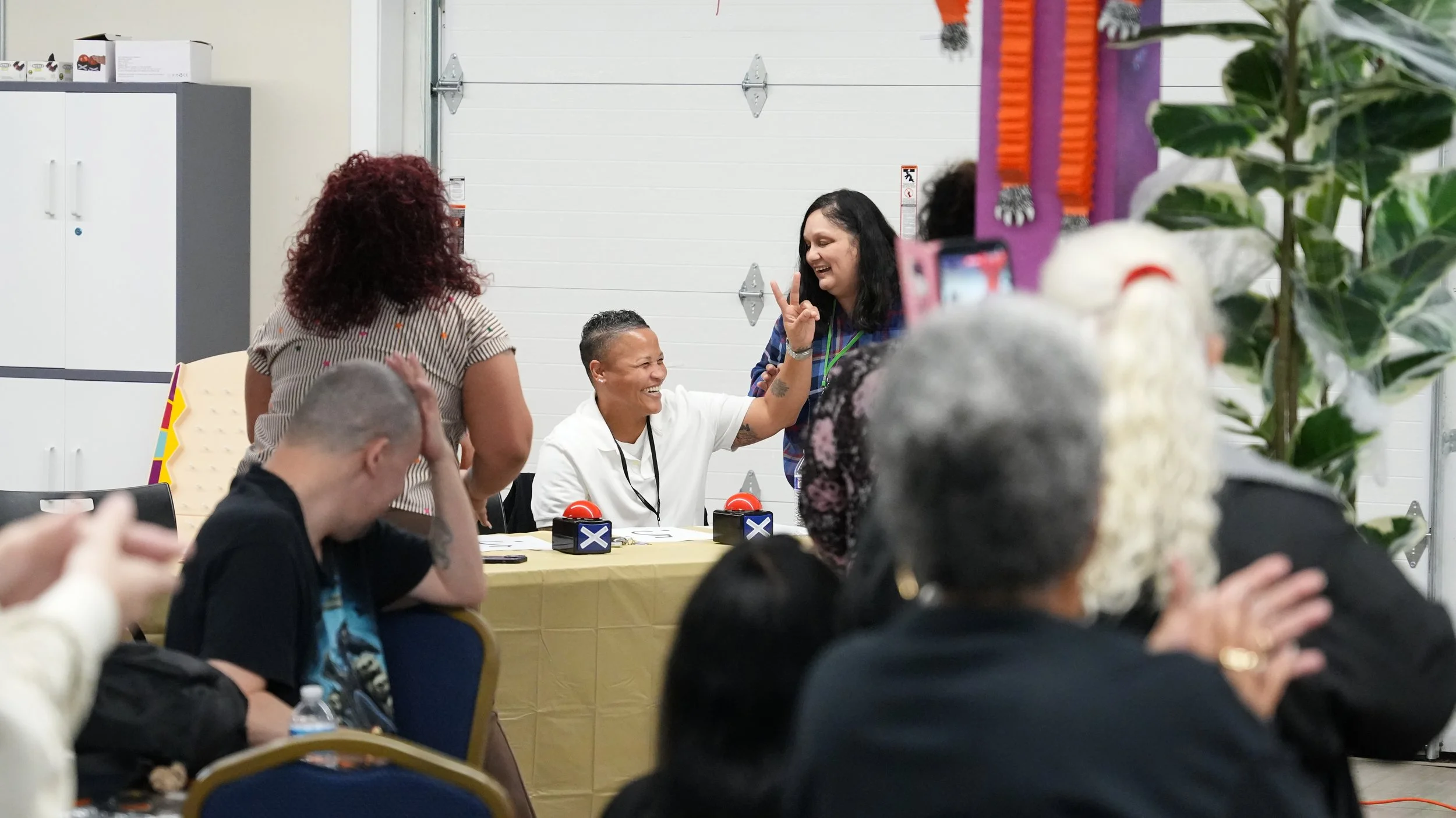 People at a panel event with a woman in white sitting at a table, smiling, daycare, making a peace sign, surrounded by others in a meeting or celebration setting.