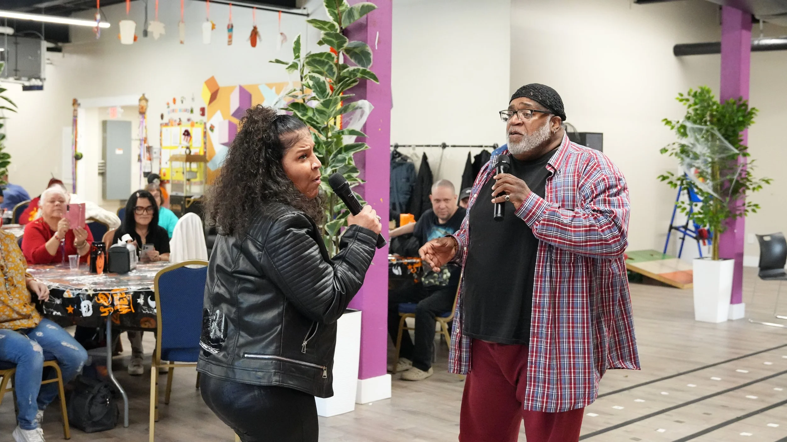 Woman and man each holding a microphone and singing or speaking at an indoor event with seated audience in the background.